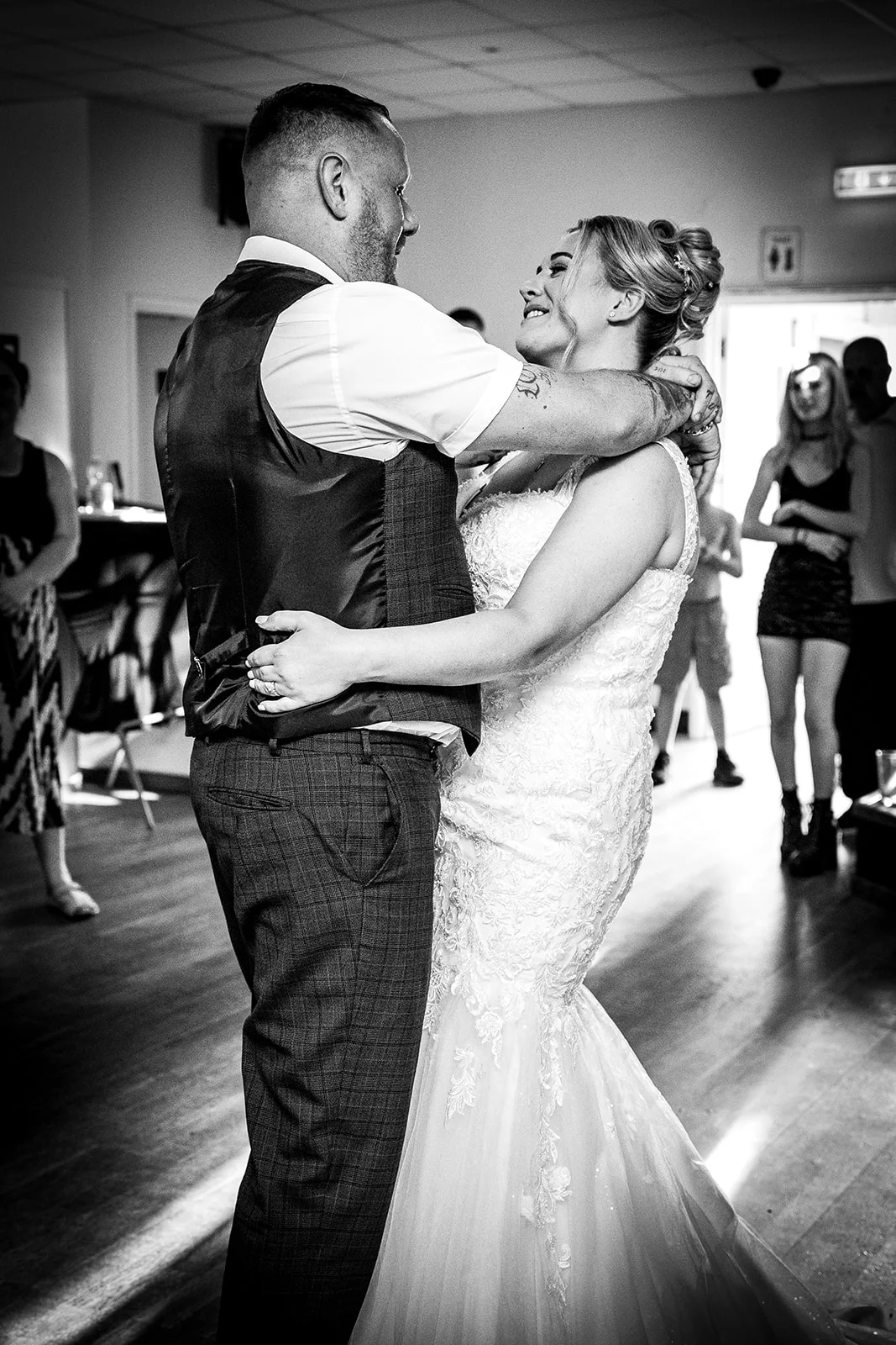 A newlywed couple shares an intimate first dance together indoors while guests look on, captured in a timeless black and white style.
