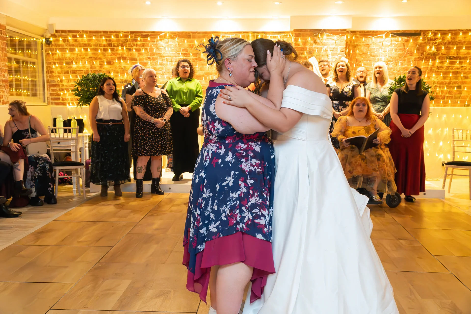 A bride in a white satin off-the-shoulder gown shares a deeply emotional embrace with a family member on a wooden dance floor, with a choir singing in the background against an exposed brick wall decorated with fairy lights.