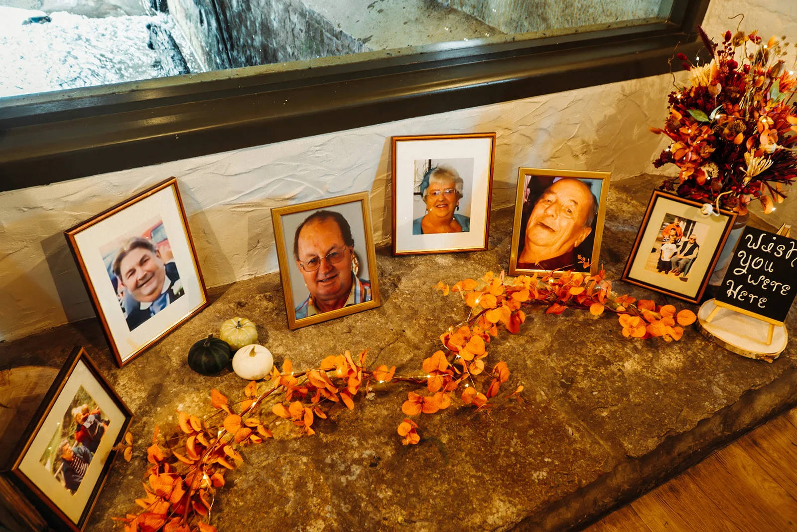 A thoughtful memory table features framed family photographs arranged alongside autumn foliage and a small chalkboard sign reading wish you were here.