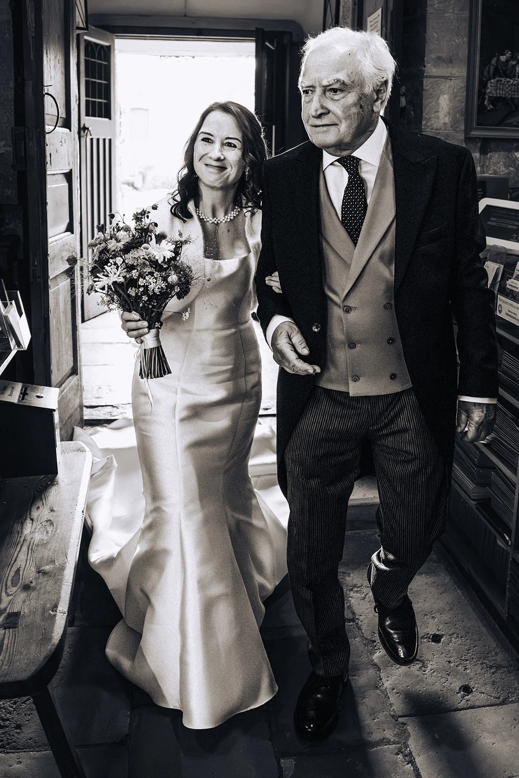 A bride smiling lovingly while walking arm in arm with her father through a doorway into a church ceremony.