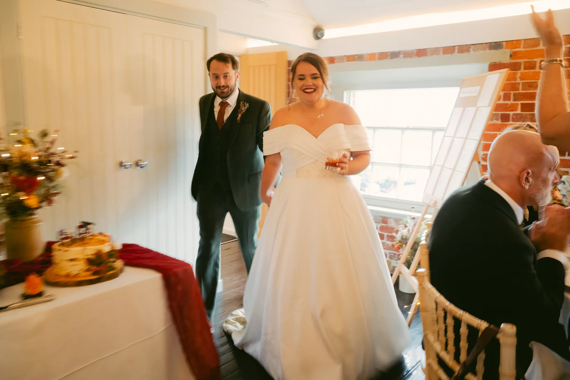 A newlywed couple joyfully enters their wedding reception in a rustic barn venue, with the smiling bride holding a glass and wearing an off-the-shoulder a-line gown while her groom follows closely in a dark green suit.