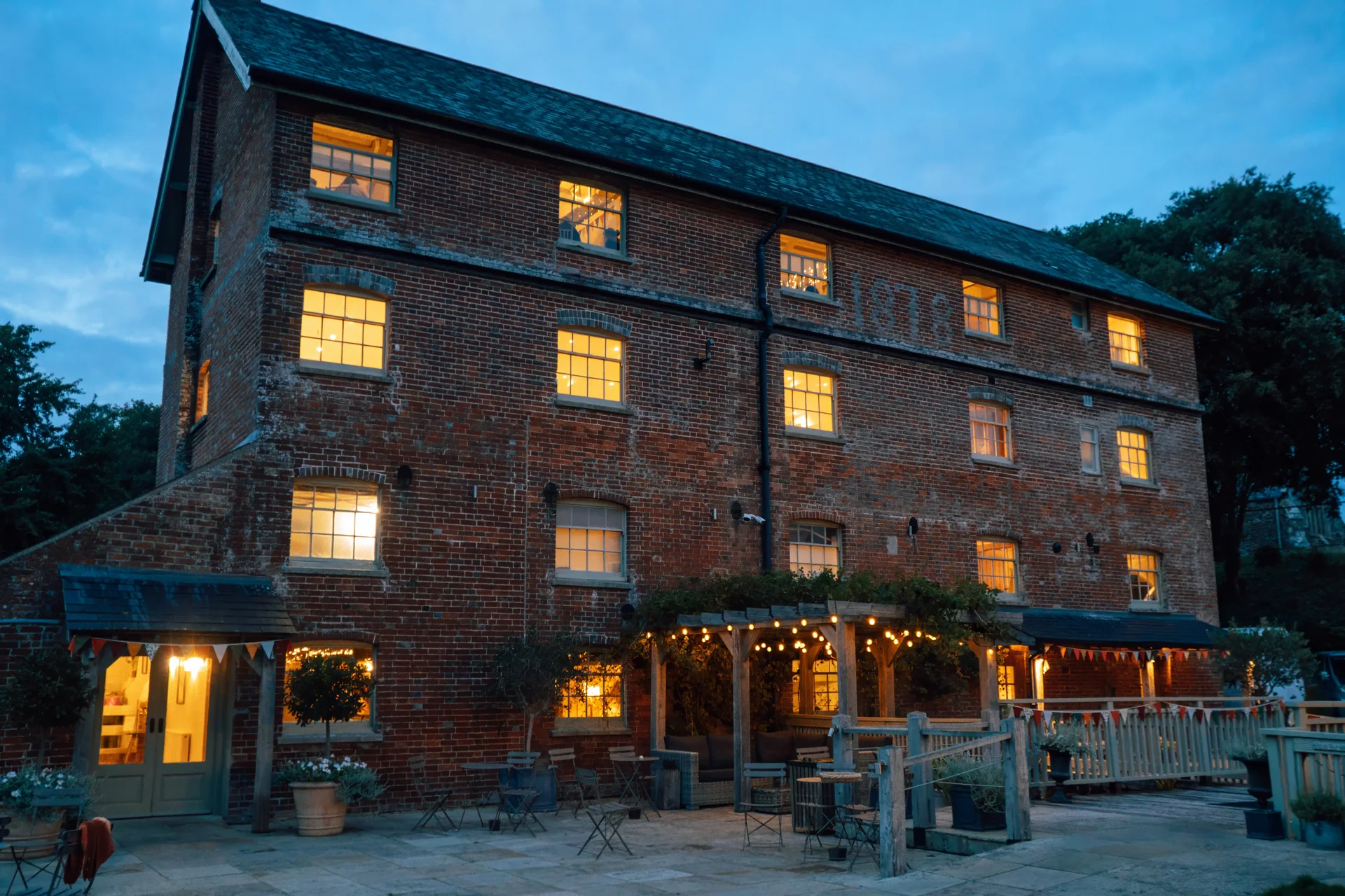 A historic red brick industrial wedding venue illuminated by warm window lights and outdoor festoon lighting during twilight.