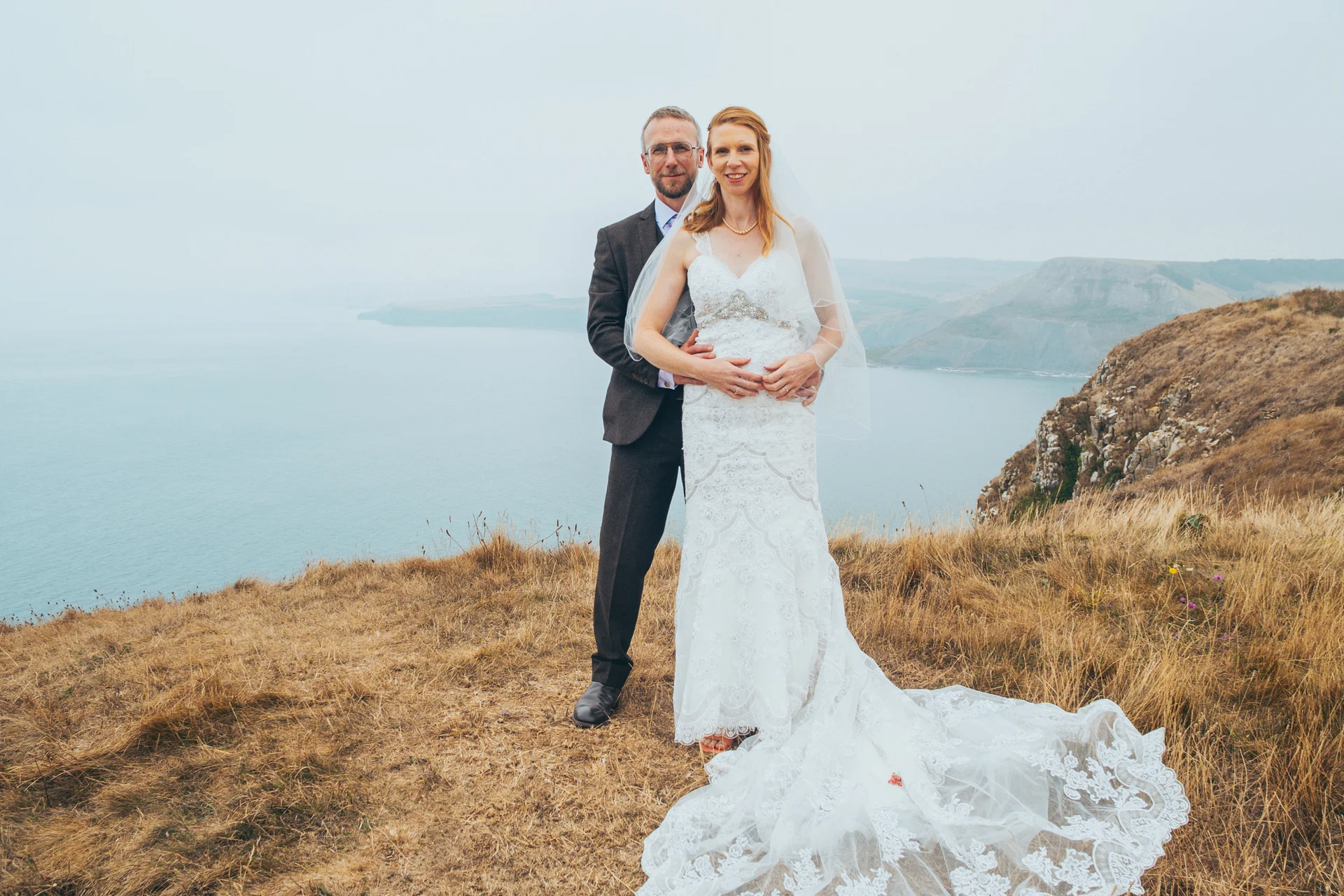 A newlywed couple poses together on a dramatic clifftop overlooking the sea on their wedding day.