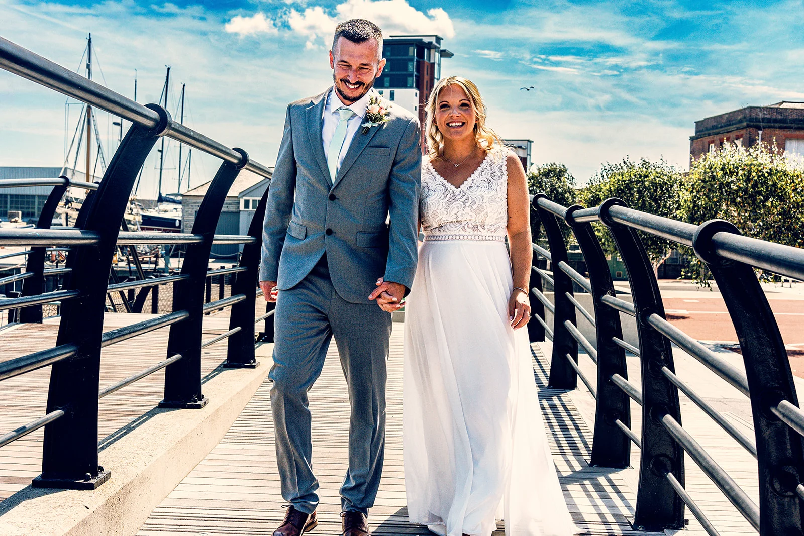 A smiling bride in a lace top wedding dress and groom in a grey suit walk hand-in-hand along a wooden boardwalk by the harbour on a sunny day.