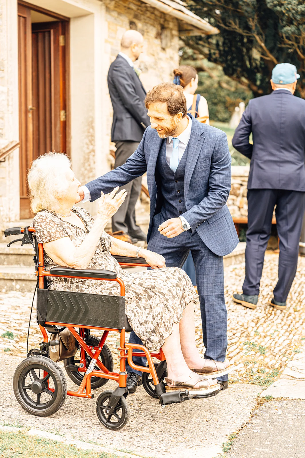 A smiling groom wearing a blue checked three-piece suit lovingly greets an elderly woman seated in a wheelchair outside a venue.