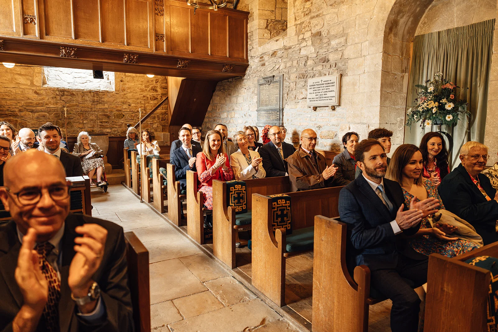 Wedding guests seated in a historic stone church clap and smile joyfully during the ceremony.