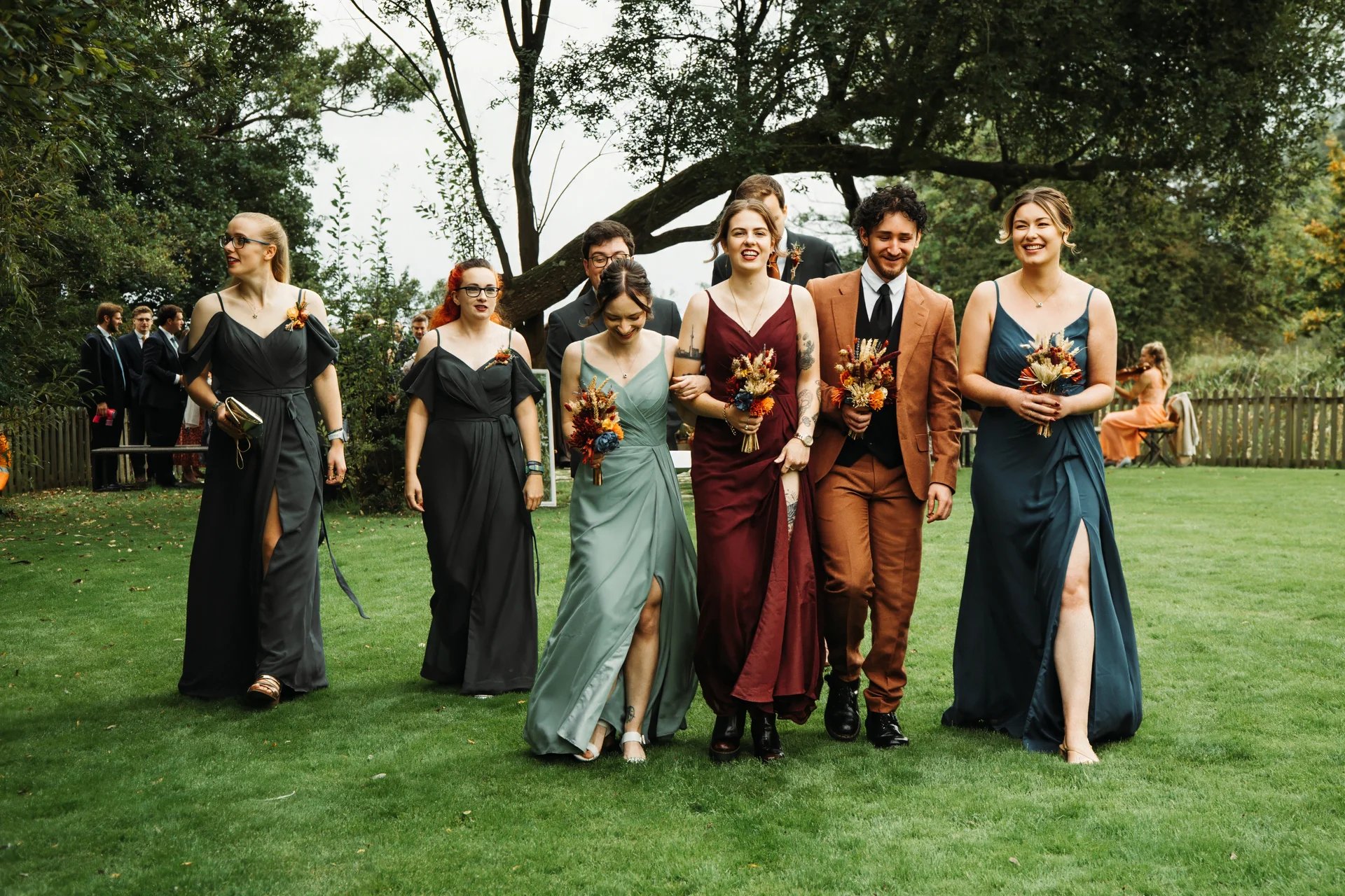 A smiling wedding party with mismatched bridesmaids in sage, burgundy, and dark grey dresses walks across a green lawn outdoors.