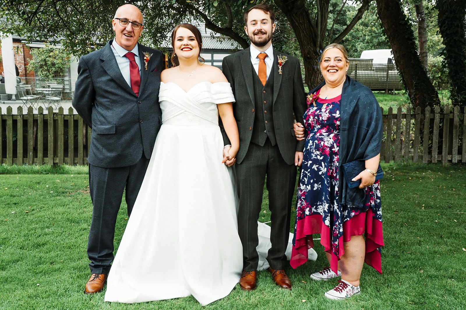 A newlywed couple poses outdoors on a lush green lawn alongside the bride's parents in front of a rustic wooden fence.