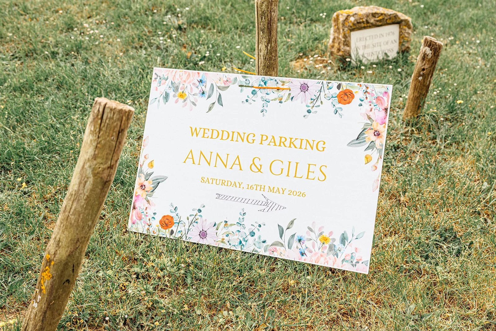 A white wedding parking sign featuring floral illustrations and gold typography stands upright in a grassy field surrounded by wooden posts.