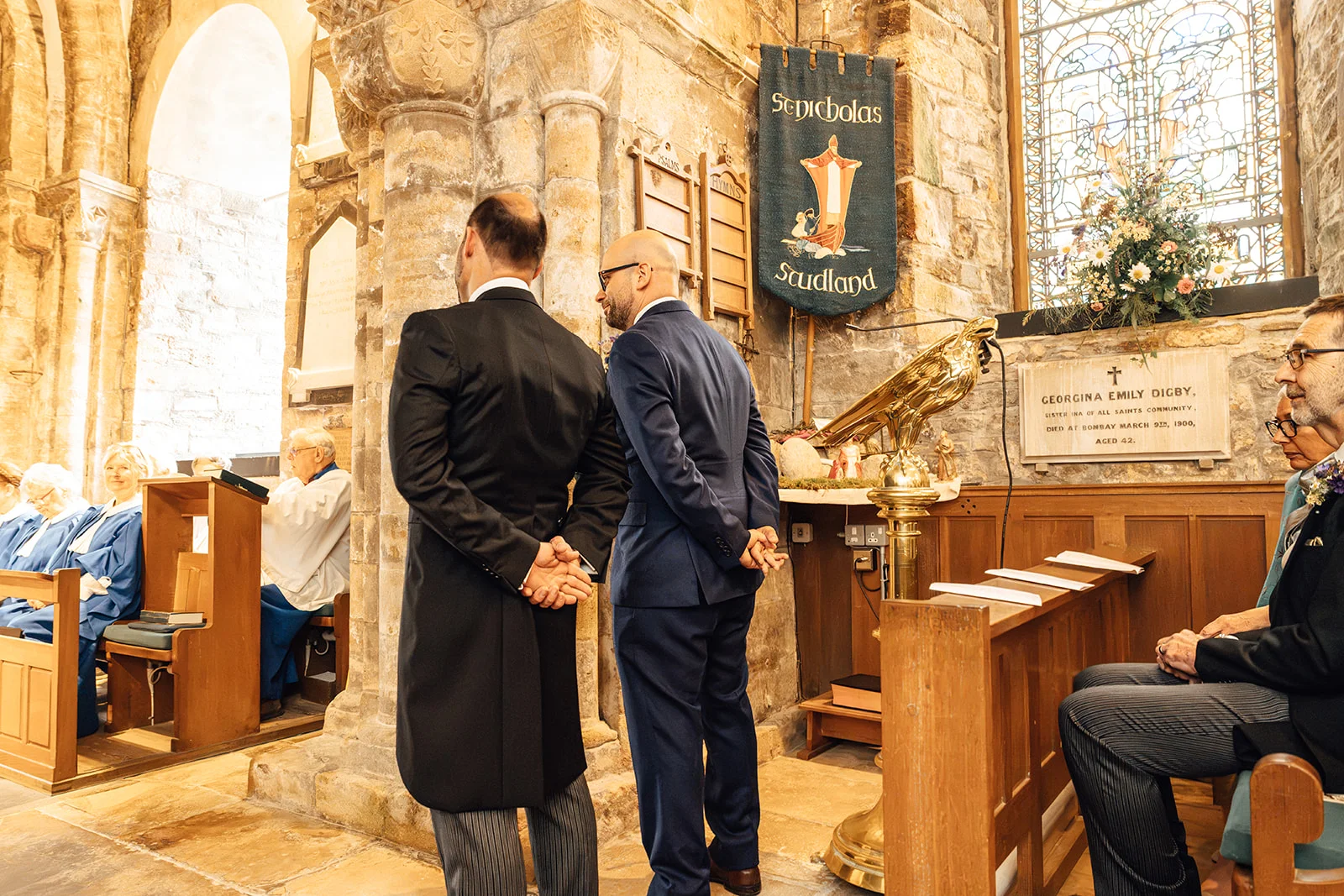 Two grooms stand inside a historic stone church during their wedding ceremony, wearing smart suits and facing the altar.