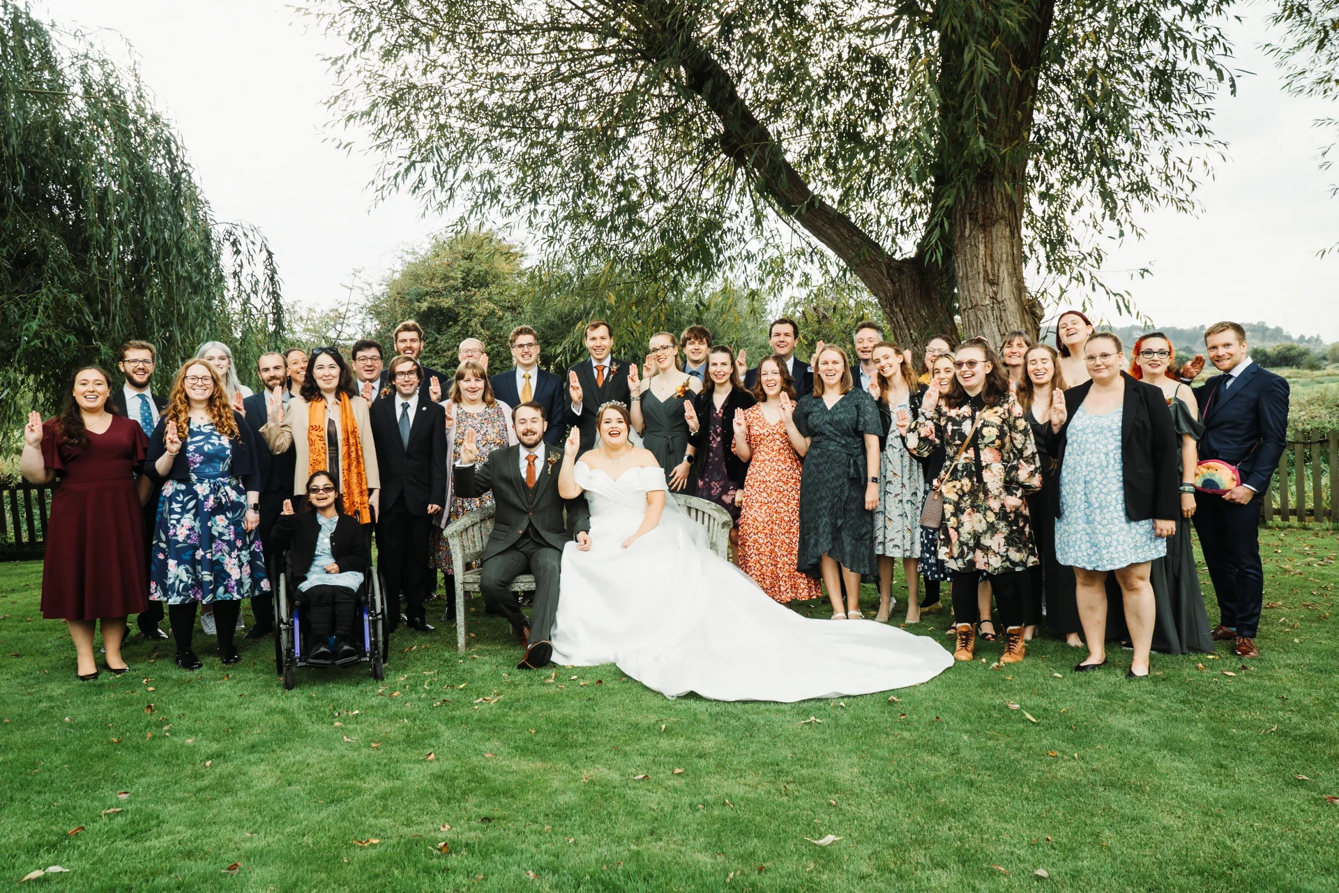 A large group of wedding guests and the newlyweds pose outdoors on a manicured lawn beneath a large tree, all holding up three fingers in a playful gesture.