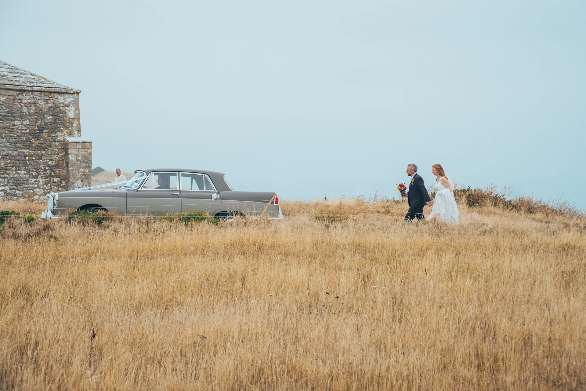 Newlyweds walk hand in hand through a tall grass field near a classic vintage car and a stone building under an overcast sky.