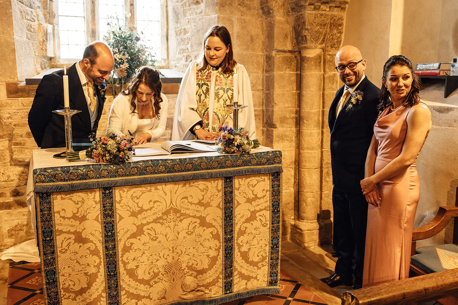 The bride and groom sign the register at an altar inside an ancient stone church while witnesses and an officiant look on smiling.