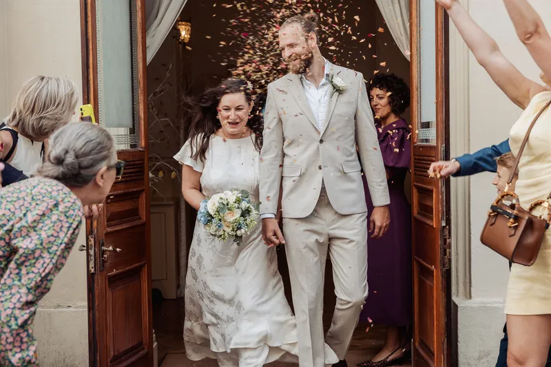 Newlyweds joyfully exit through wooden double doors while guests shower them with a cascade of colourful confetti.