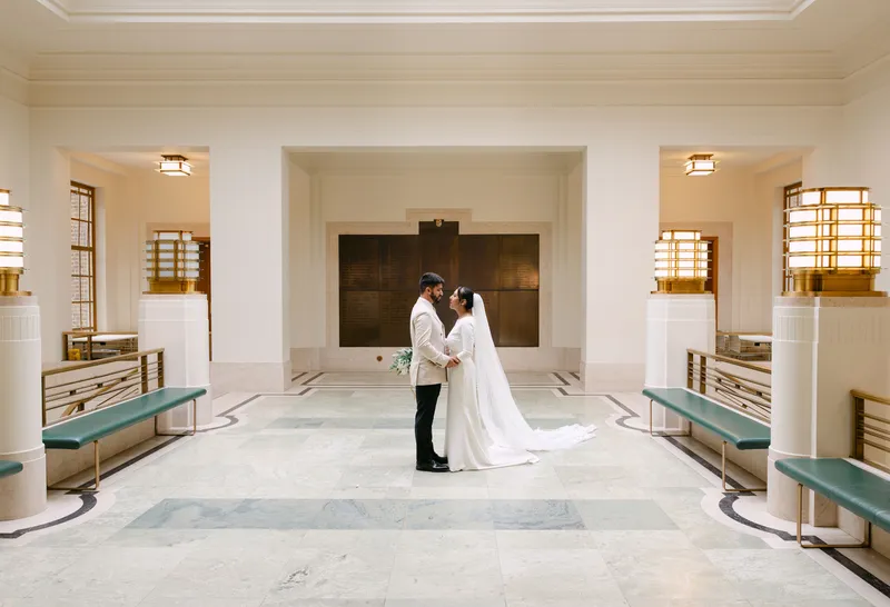A bride in a sleek crepe gown with a chapel veil and a groom in a cream suit stand nose-to-nose in a grand, symmetrical hall.