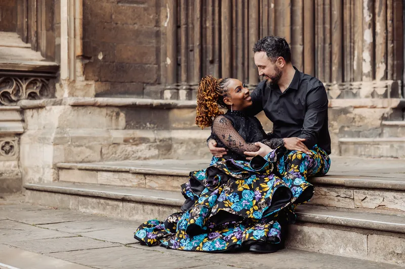 An interracial newlywed couple sitting lovingly on stone steps outside a historic building, with the bride wearing a striking black floral print gown with a ruffled skirt.
