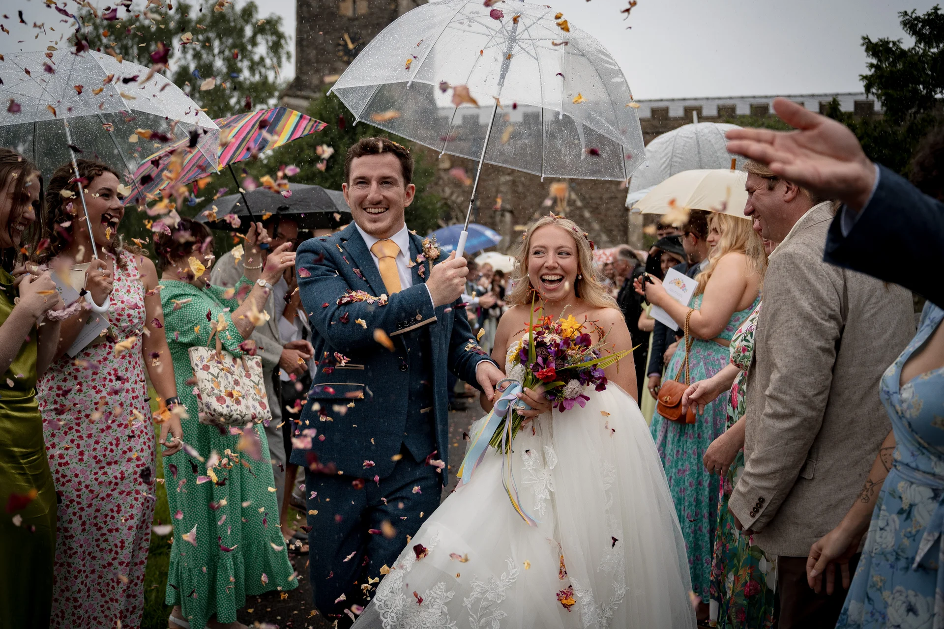 A joyful bride and groom laugh and walk through a shower of colourful confetti outside a church on a rainy wedding day, holding clear umbrellas to shield themselves.