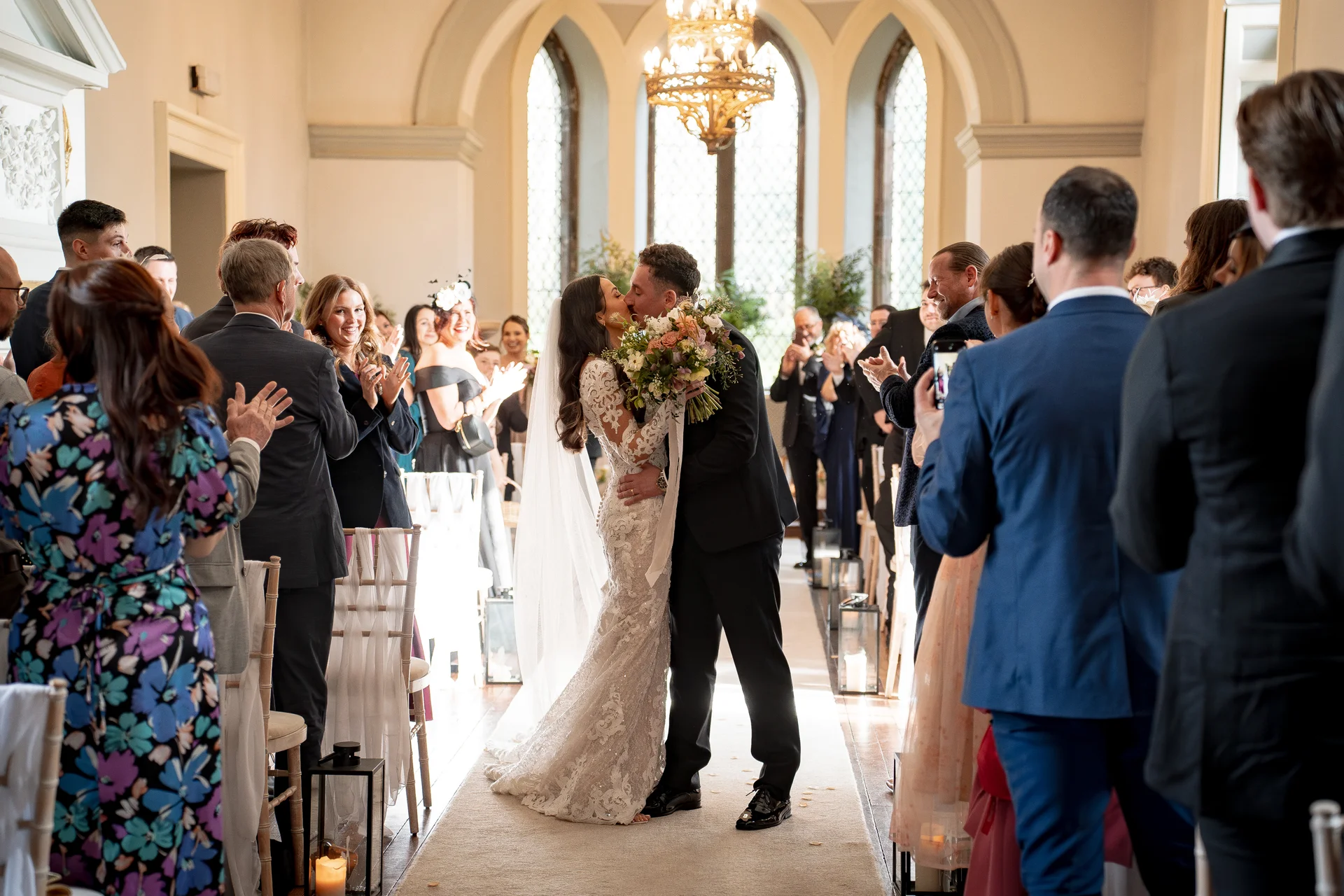 The newlyweds share a tender first kiss as they walk down the aisle surrounded by cheering guests in a sunlit historic venue.