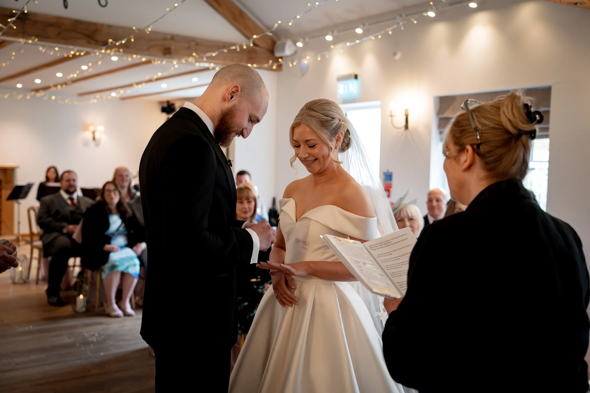 The groom places a ring on the bride's finger during their indoor wedding ceremony while a celebrant holds a service book nearby.