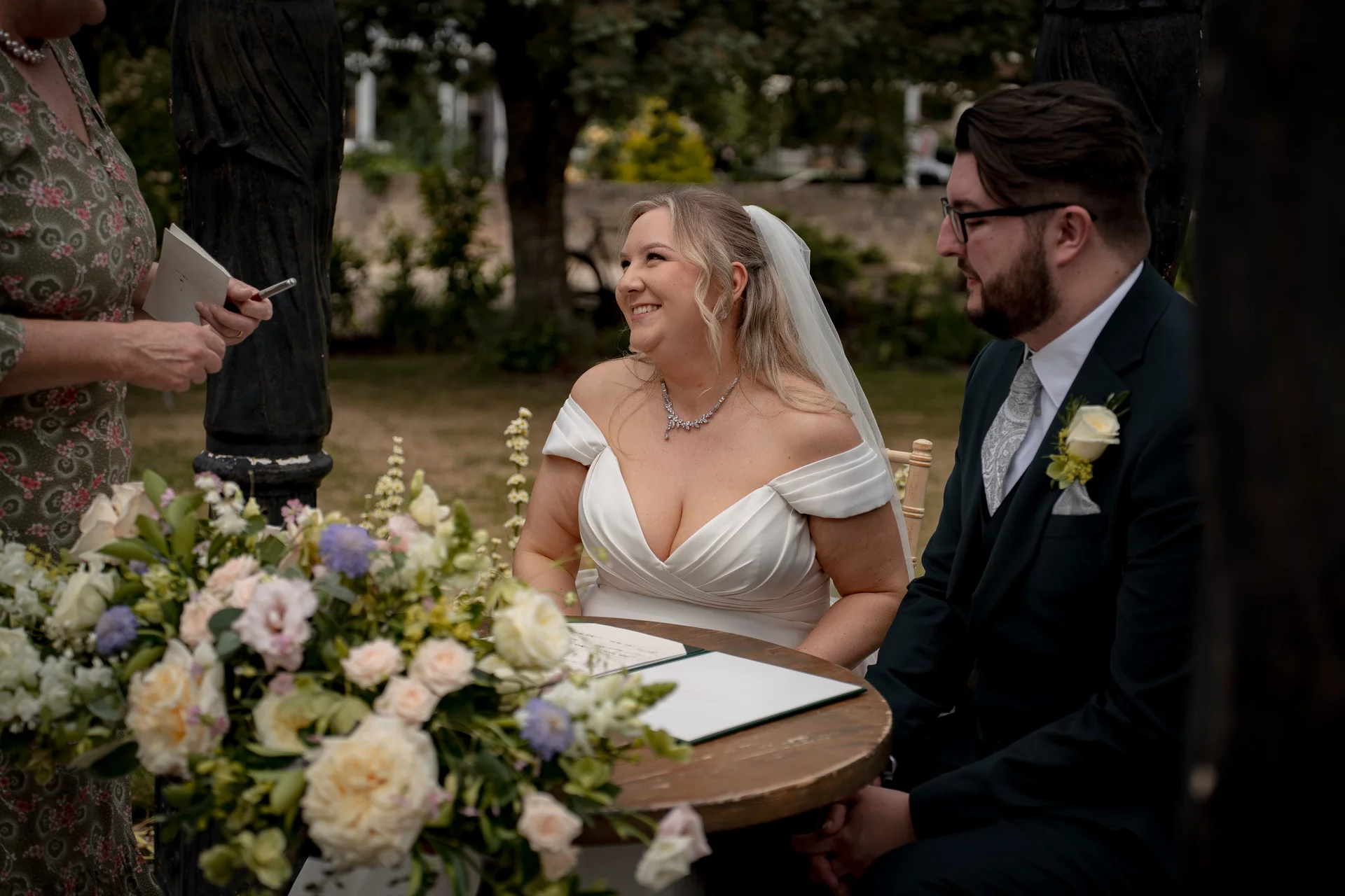 The bride and groom smile joyfully during the register signing at their outdoor celebrant-led ceremony, seated beside a lush floral arrangement.