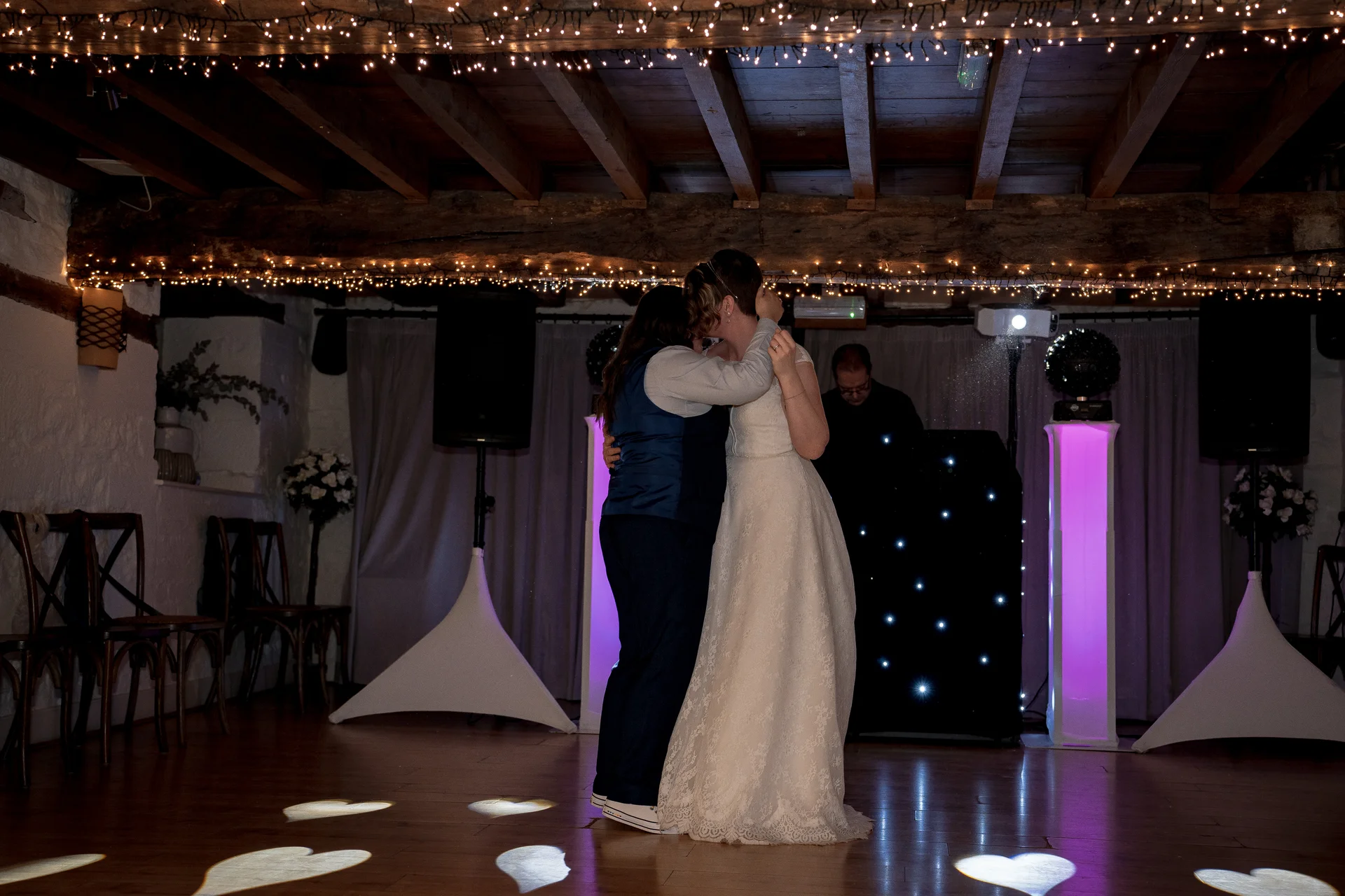 Two brides share their first dance in a rustic barn venue decorated with exposed wooden beams and glowing fairy lights above.