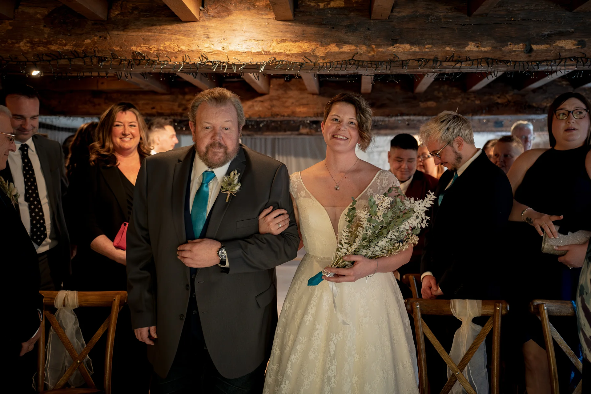 A smiling short-haired bride walks down the aisle arm in arm with her father inside a rustic barn venue with exposed wooden beams.