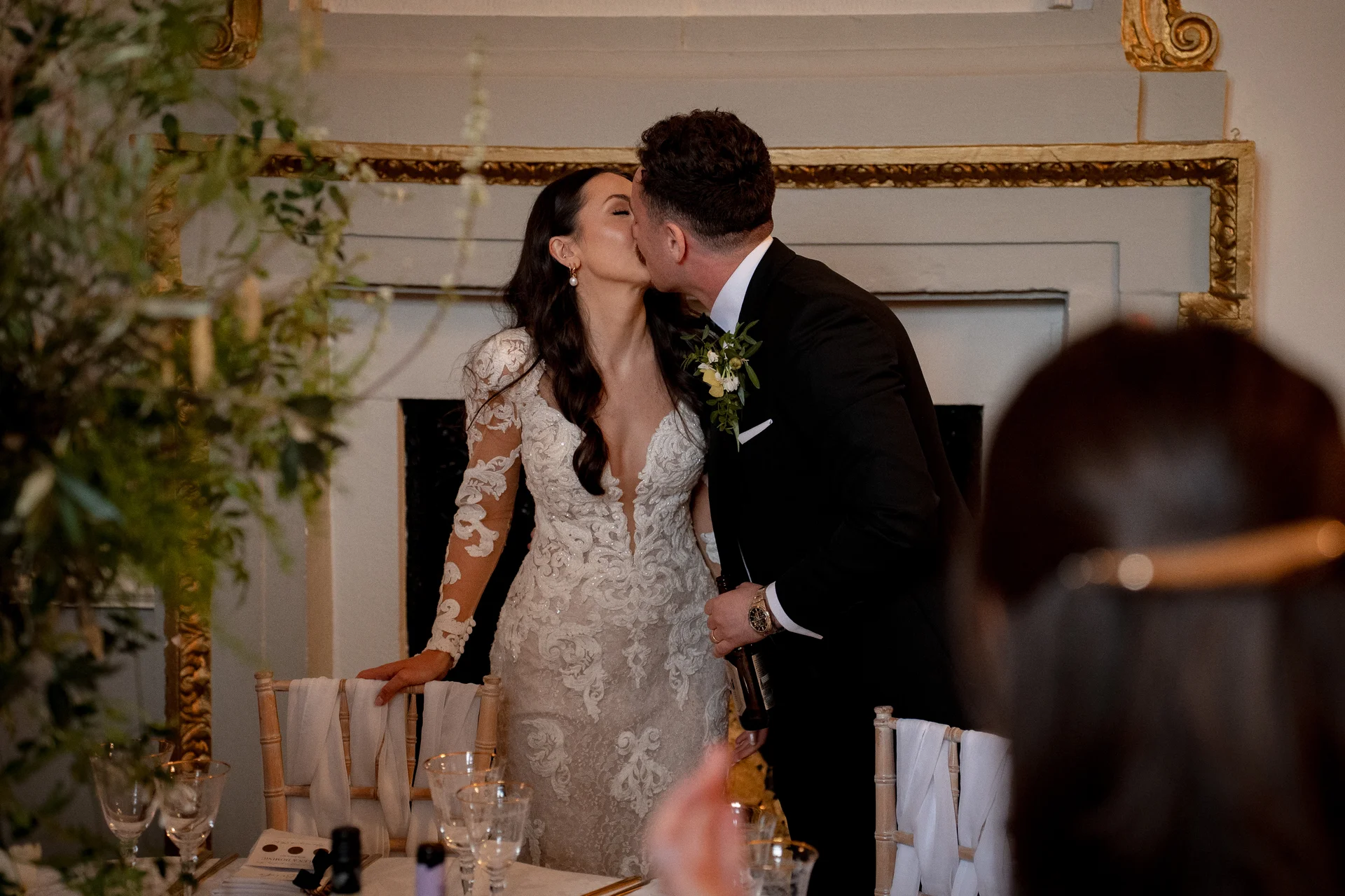 A newlywed couple shares a tender kiss indoors near an elegant fireplace while seated at the top table during their wedding reception.