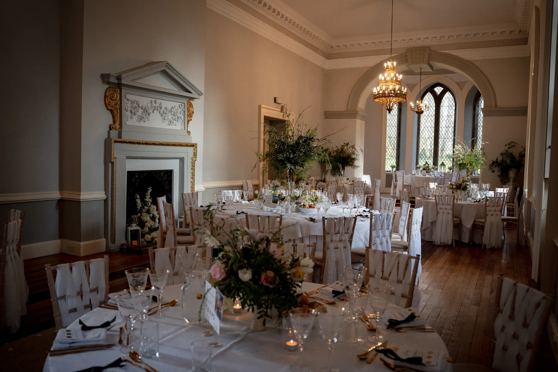 An elegant wedding reception room featuring round tables set with white linens, floral centrepieces, glowing candles, and crystal chandeliers hanging from the high ceiling.