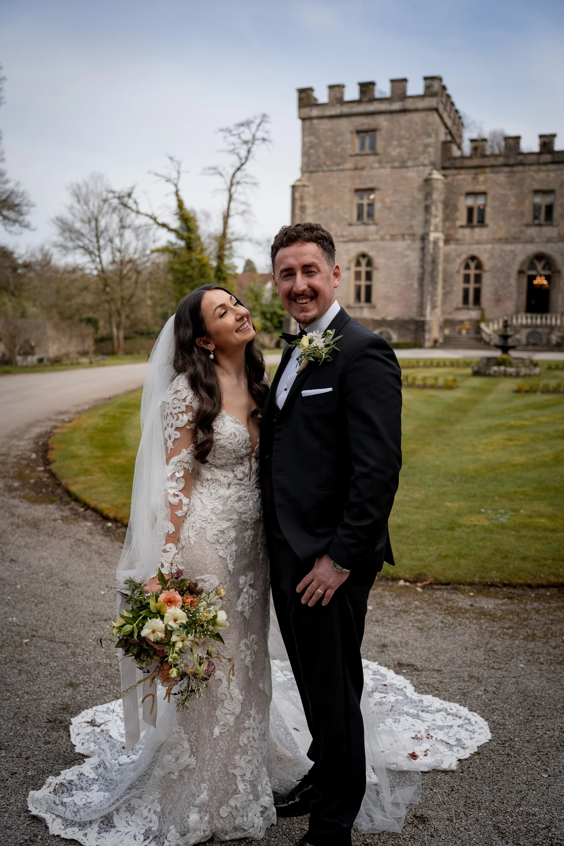A smiling bride and groom pose outside a historic stone castle venue, with the bride holding a textured bouquet and wearing a lace long-sleeved gown.