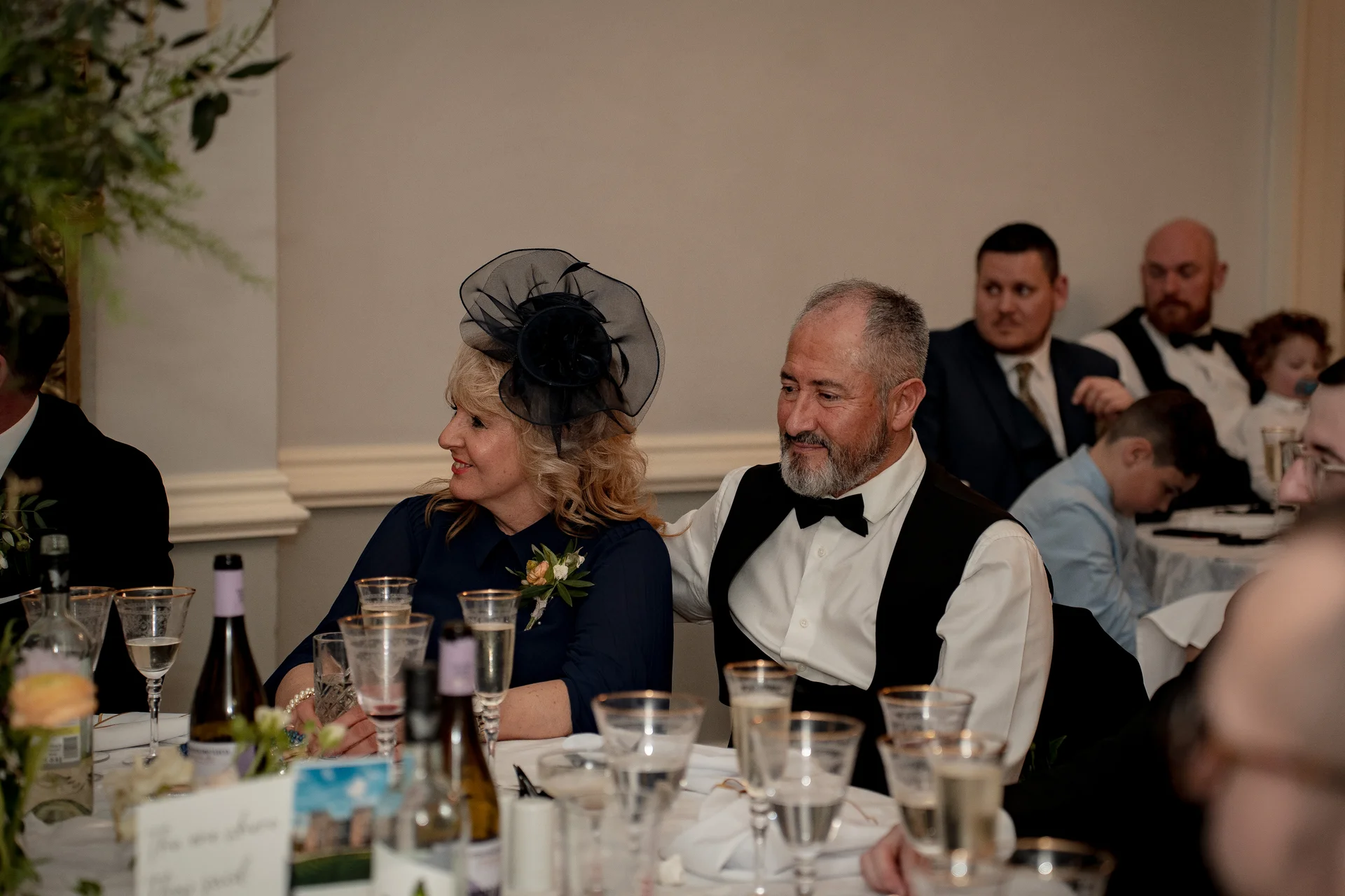 Guests laugh and smile during wedding breakfast speeches while seated at a table adorned with wine glasses and floral details.