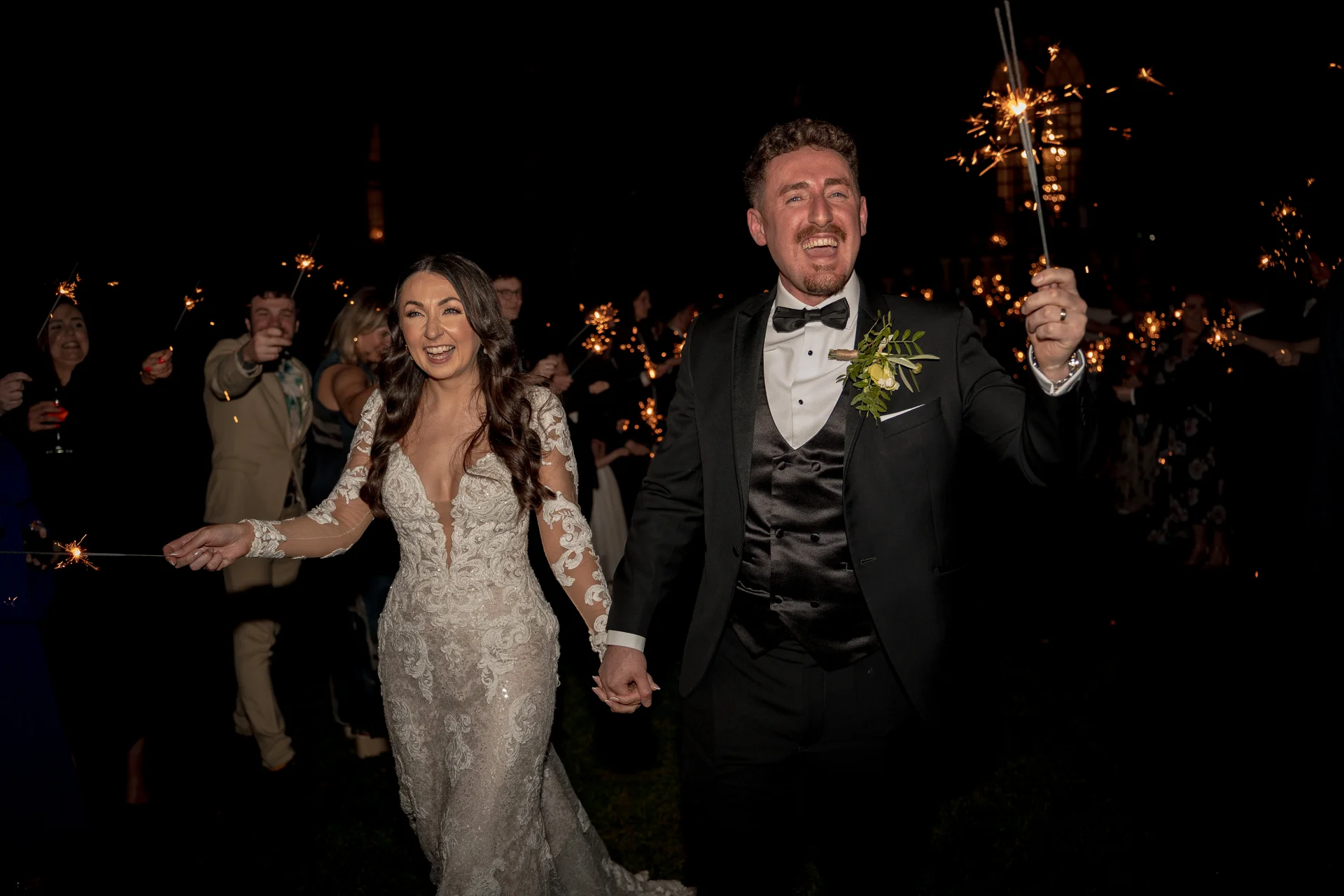 A newlywed couple joyfully walks through a glowing sparkler tunnel at night, holding hands and laughing as guests cheer them on in the background.