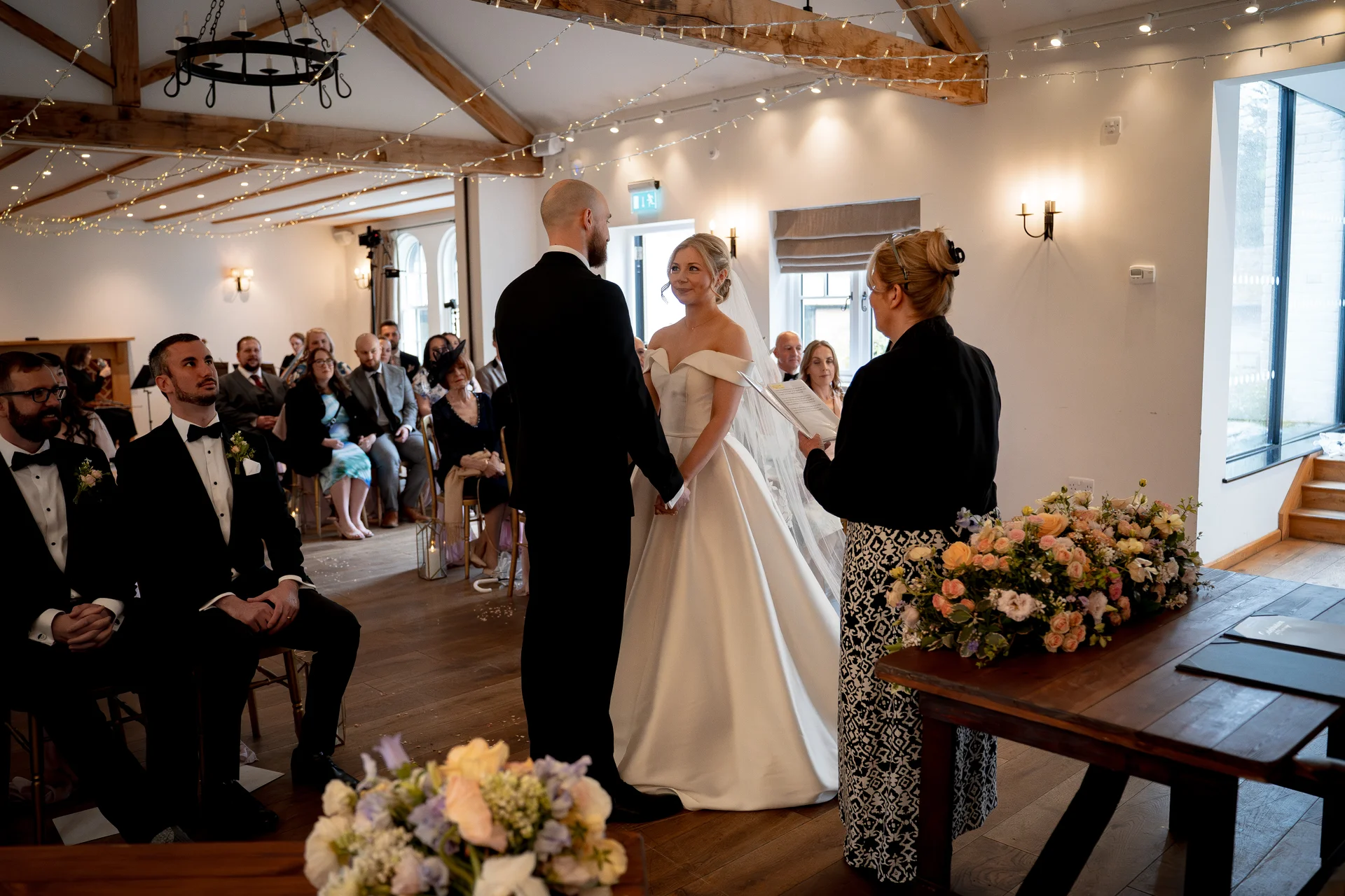 A bride and groom stand facing each other at the altar during a ceremony inside a bright barn venue decorated with exposed wooden beams and fairy lights.