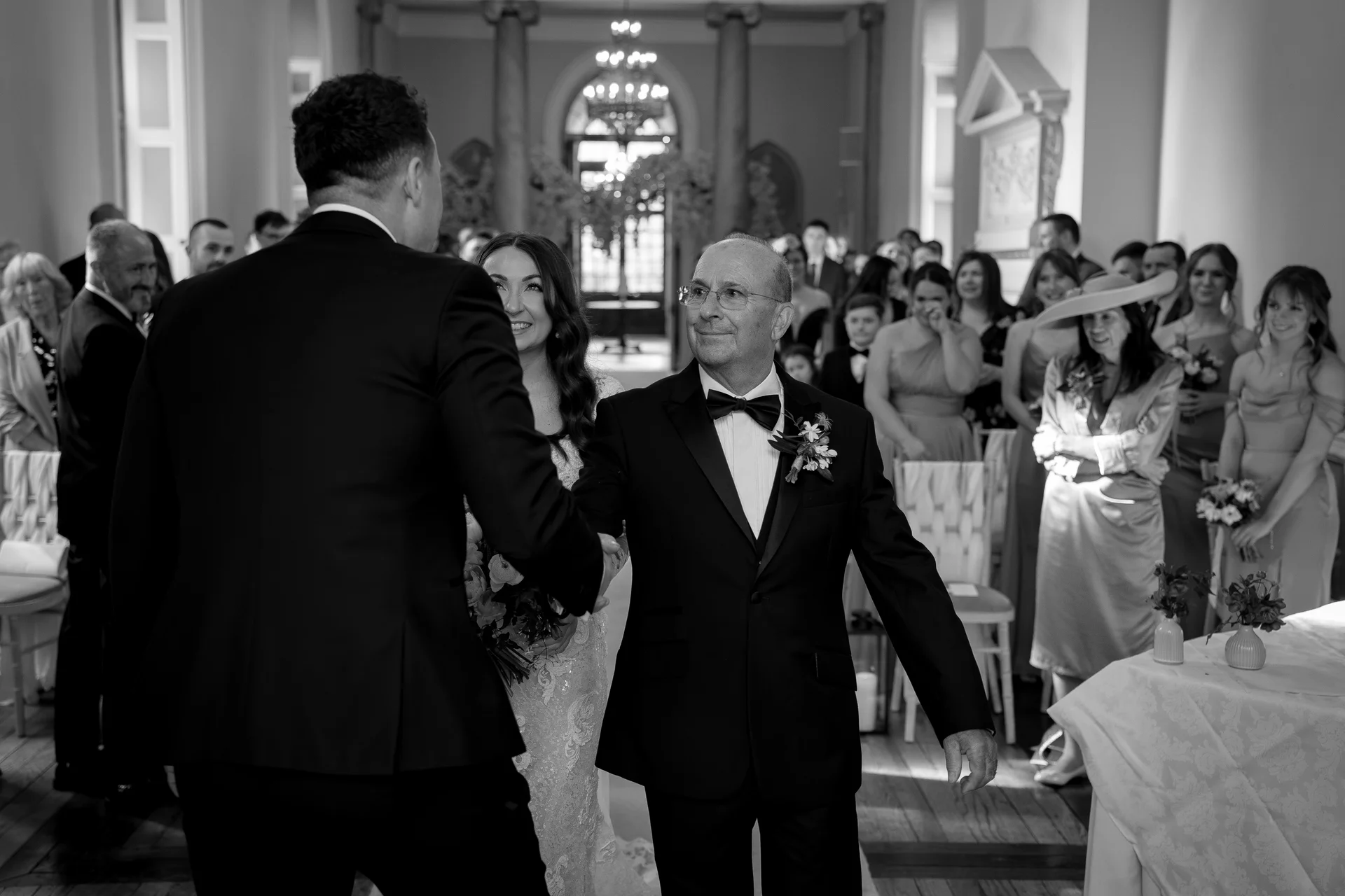 A smiling father of the bride proudly hands his daughter over to the groom at the front of a grand ceremony room.