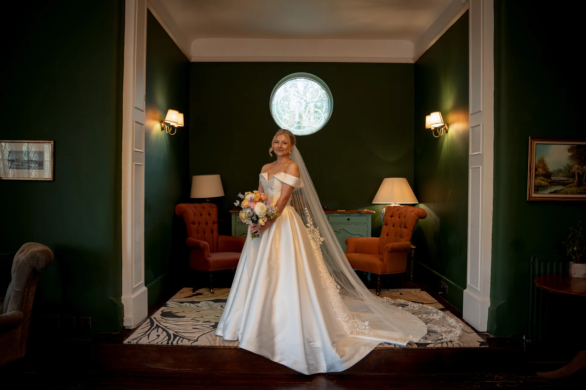 A smiling bride in an off-the-shoulder satin gown and long lace-edged veil stands holding a bouquet inside a green-panelled room framed by a doorway.