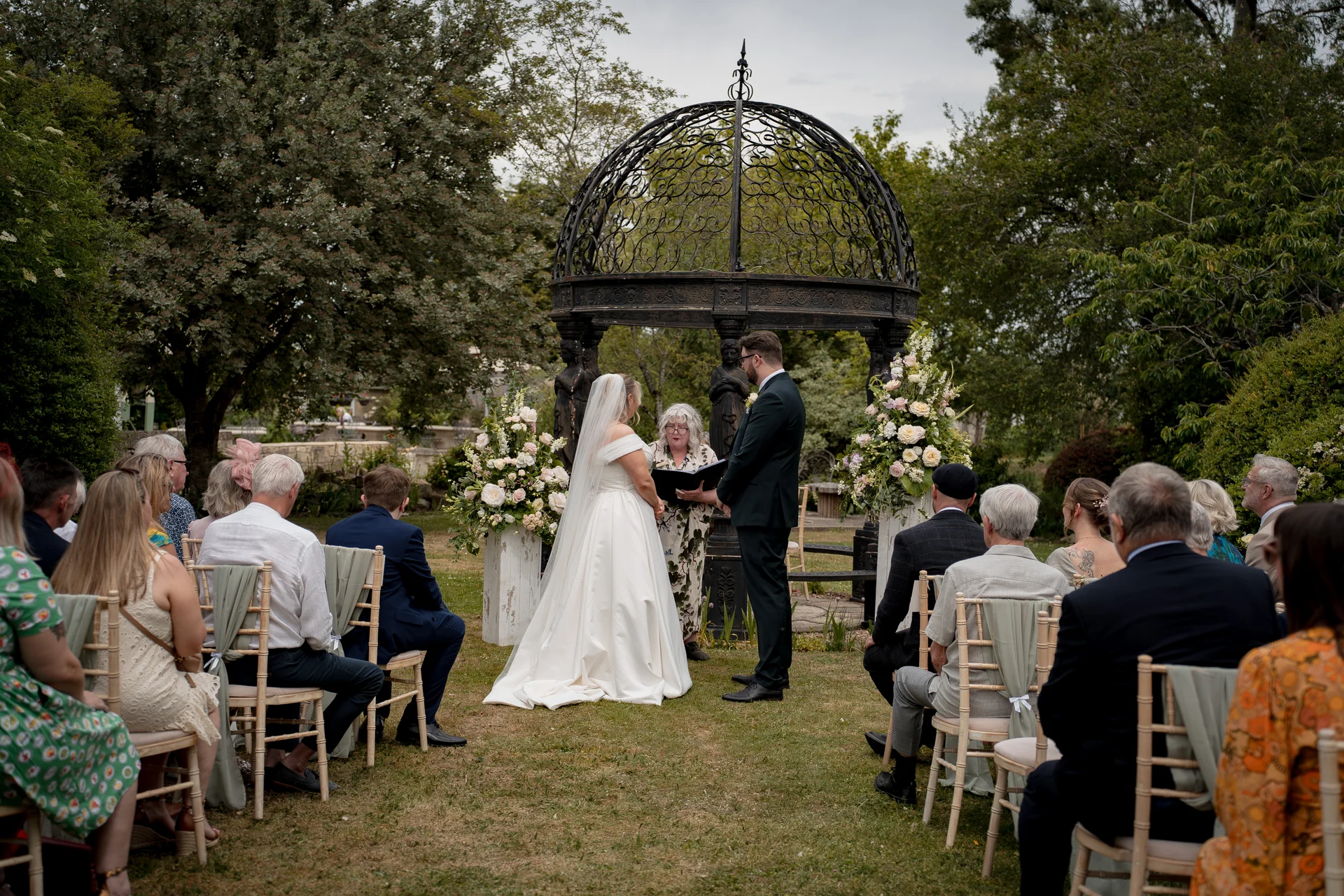 A bride and groom exchange vows during an outdoor ceremony beneath a black metal gazebo surrounded by guests seated on chairs.