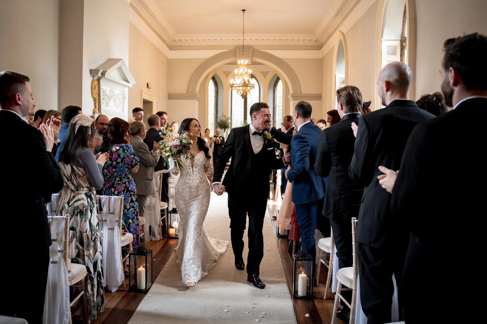 Newlywed bride and groom walk hand in hand down the aisle smiling joyfully as guests applaud and celebrate their marriage.