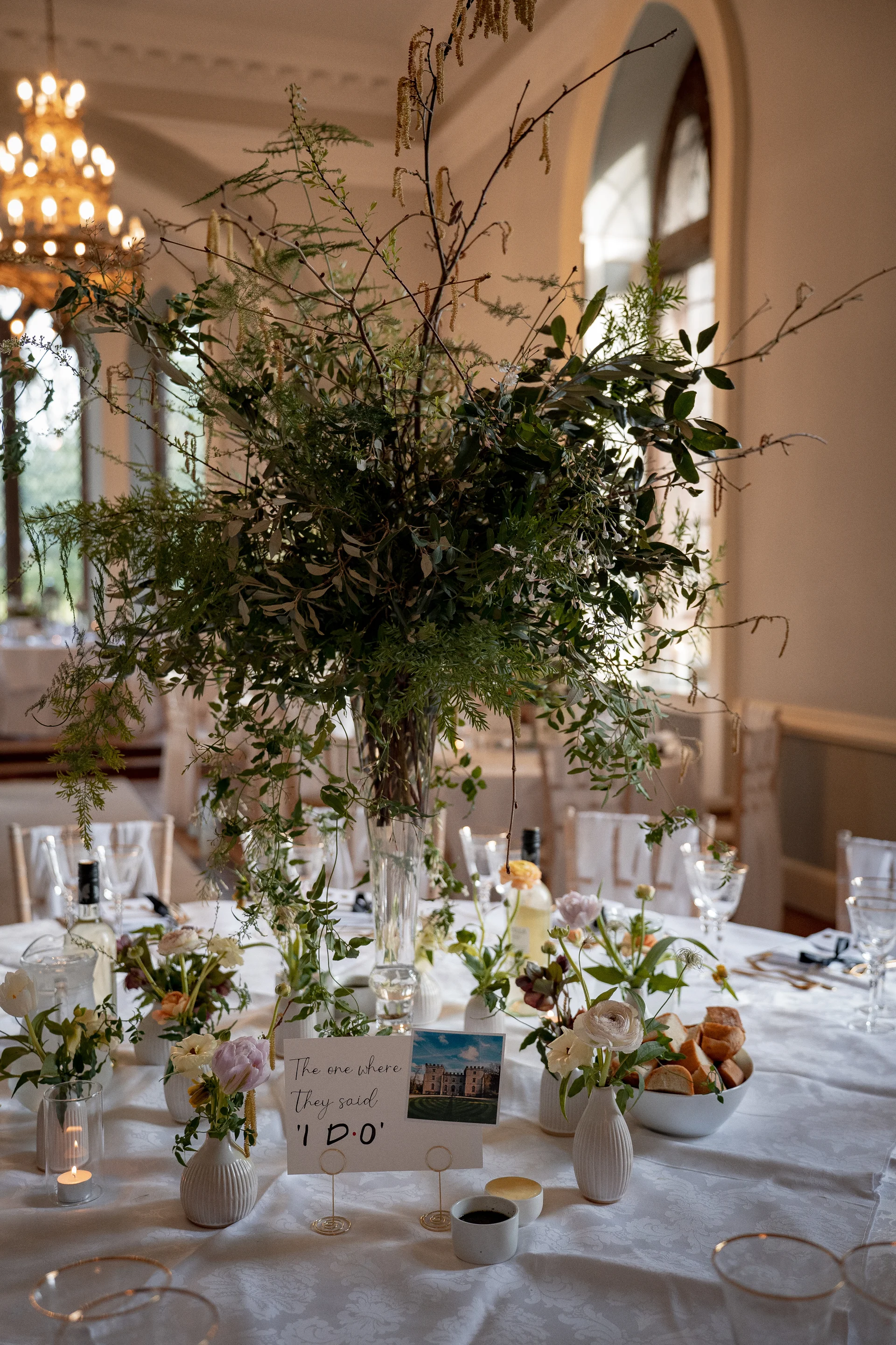 A stunning tall botanical centrepiece featuring wild foliage and branches sits proudly on a round reception table surrounded by bud vases, candles, and a custom table sign.