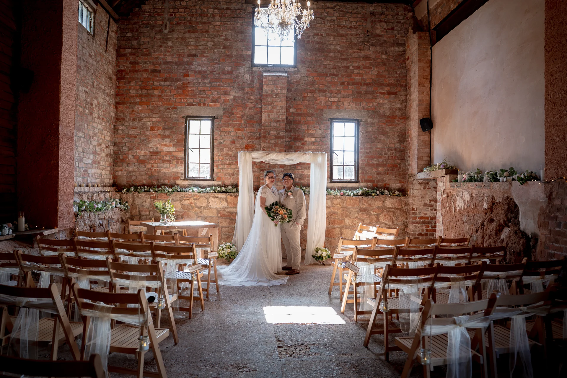 A same-sex couple stands beneath a draped ceremony arch inside a rustic barn venue featuring exposed brick walls and rows of wooden chairs.