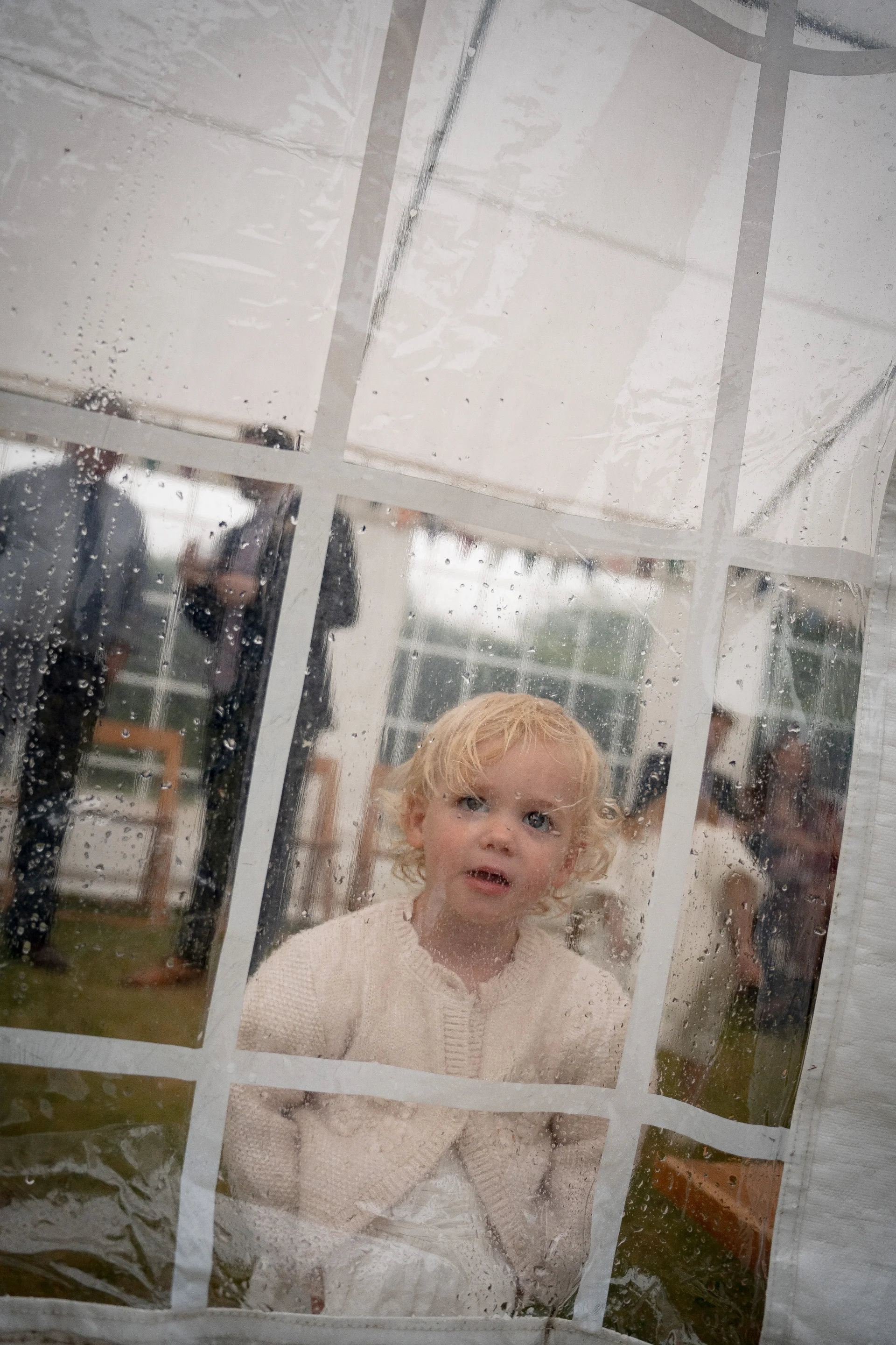 A young blonde child looks through a wet, rain-spattered clear tent window during an outdoor wedding celebration.