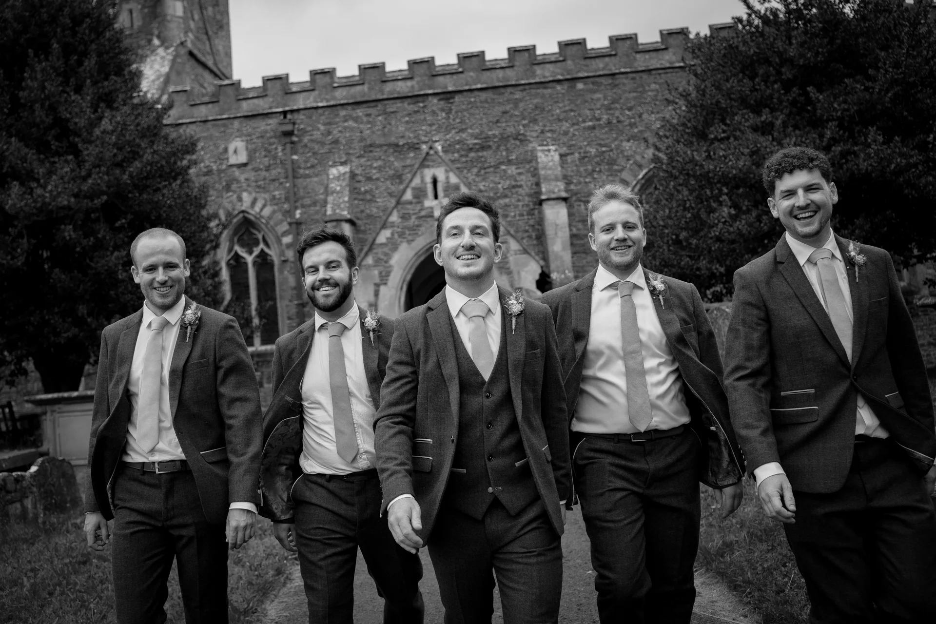 A smiling group of groomsmen in matching tweed suits walk together outside a historic stone church on the wedding day.