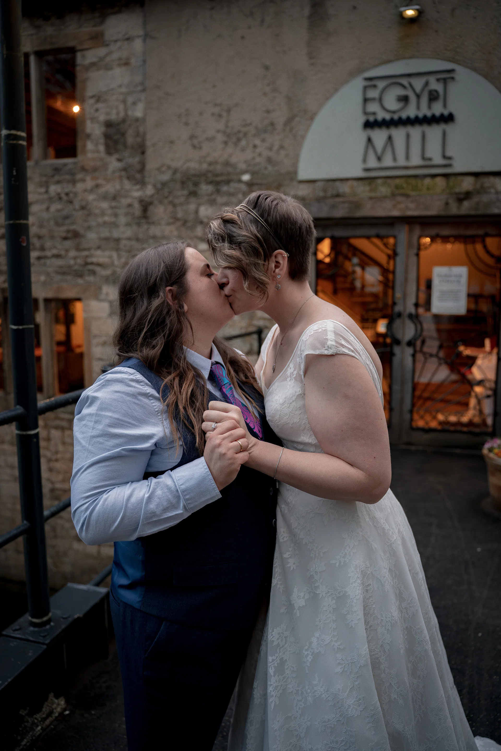 Two brides share a tender kiss outside the historic stone entrance of Egypt Mill wearing a lace wedding dress and a tailored waistcoat with a patterned tie.