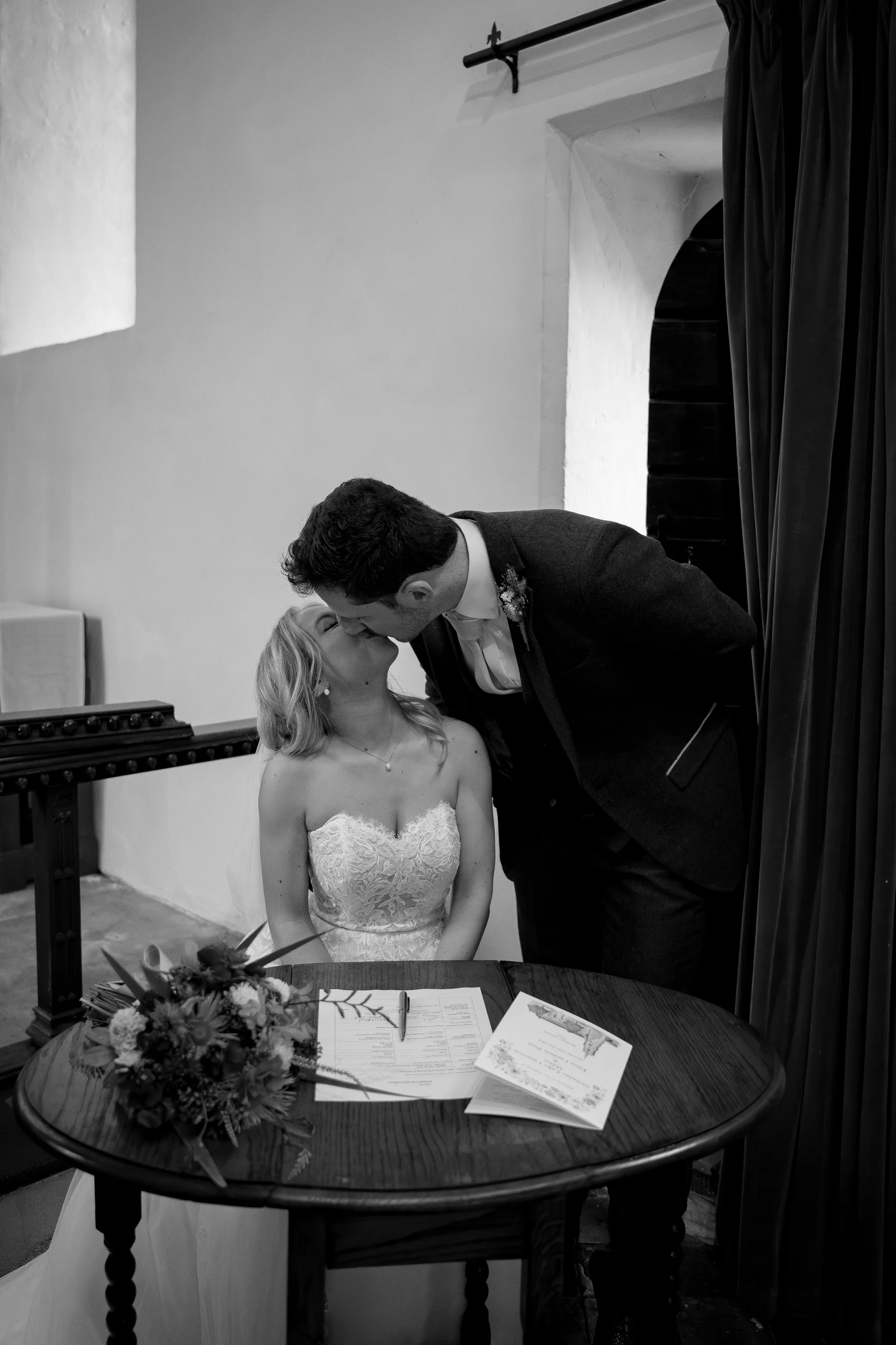 A newlywed couple share a tender kiss over the signing table following their church ceremony, with the bride in a lace sweetheart gown and the groom in a dark suit.