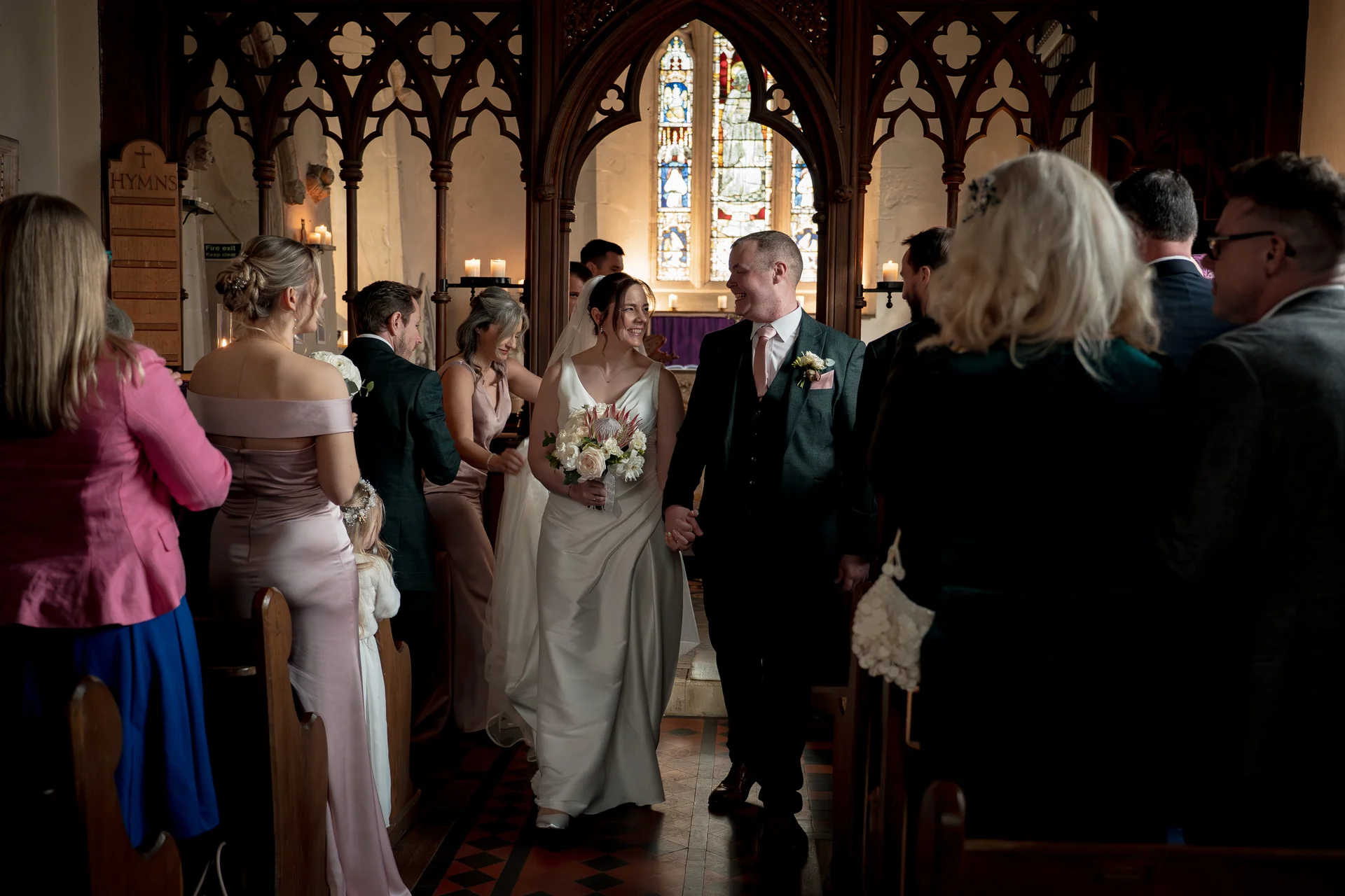Newlywed bride and groom smiling at each other while walking hand in hand down the aisle of a historic church following their ceremony.