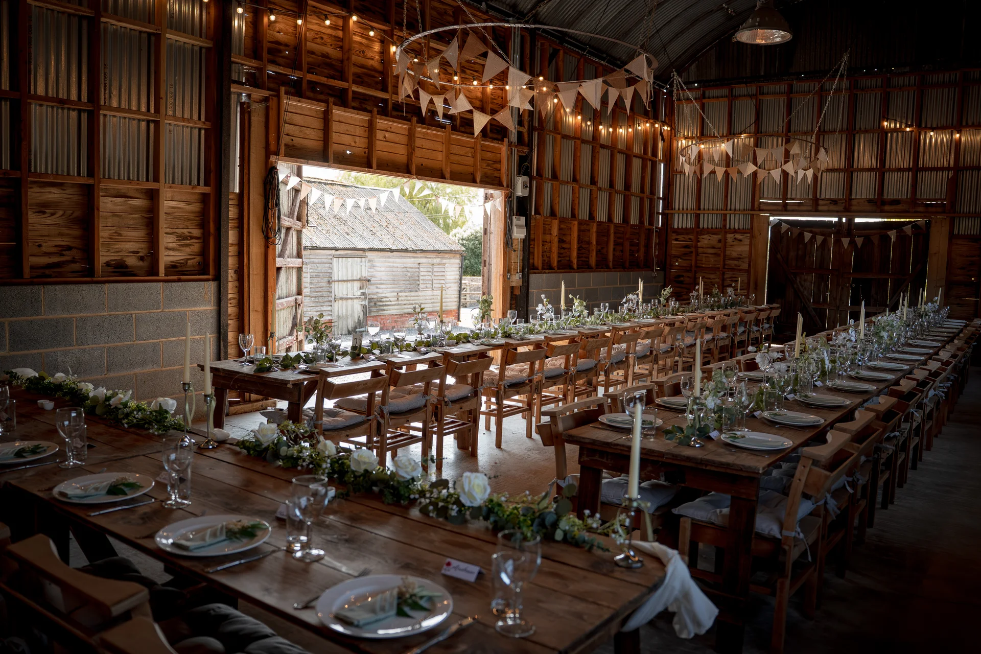 A rustic barn wedding reception featuring long wooden banquet tables set with white linens, elegant floral runners, and glowing taper candles beneath exposed wooden rafters and hanging bunting.