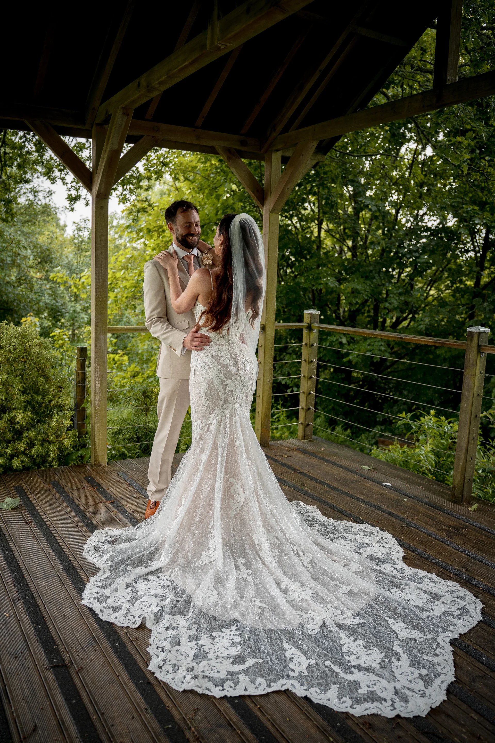 A newlywed couple embraces inside a wooden gazebo with a sweeping lace bridal train spread across the decking.