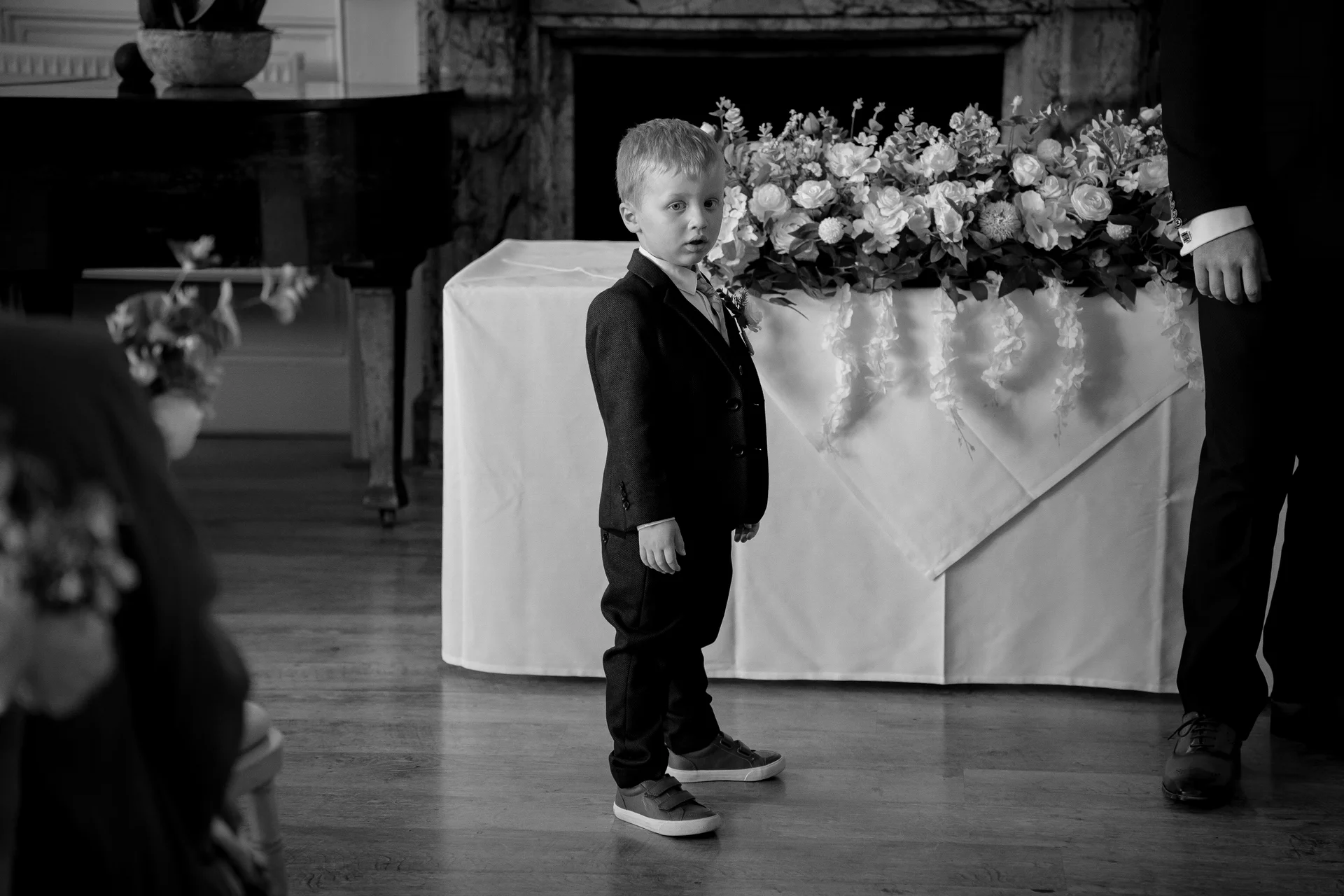A young page boy stands near a decorated table during a wedding ceremony, captured in a candid black and white shot.