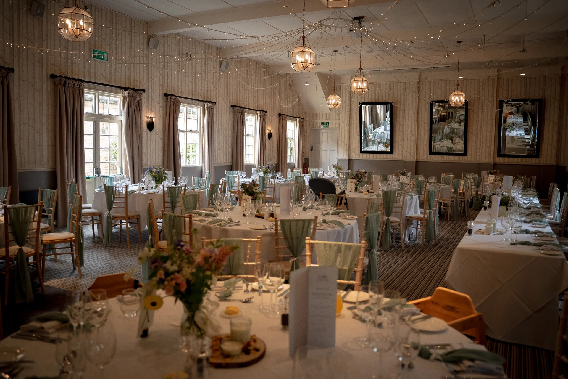 An elegantly decorated wedding reception room featuring round and long tables set with crisp white linens, sage green accents, floral centrepieces, and a canopy of twinkling fairy lights overhead.