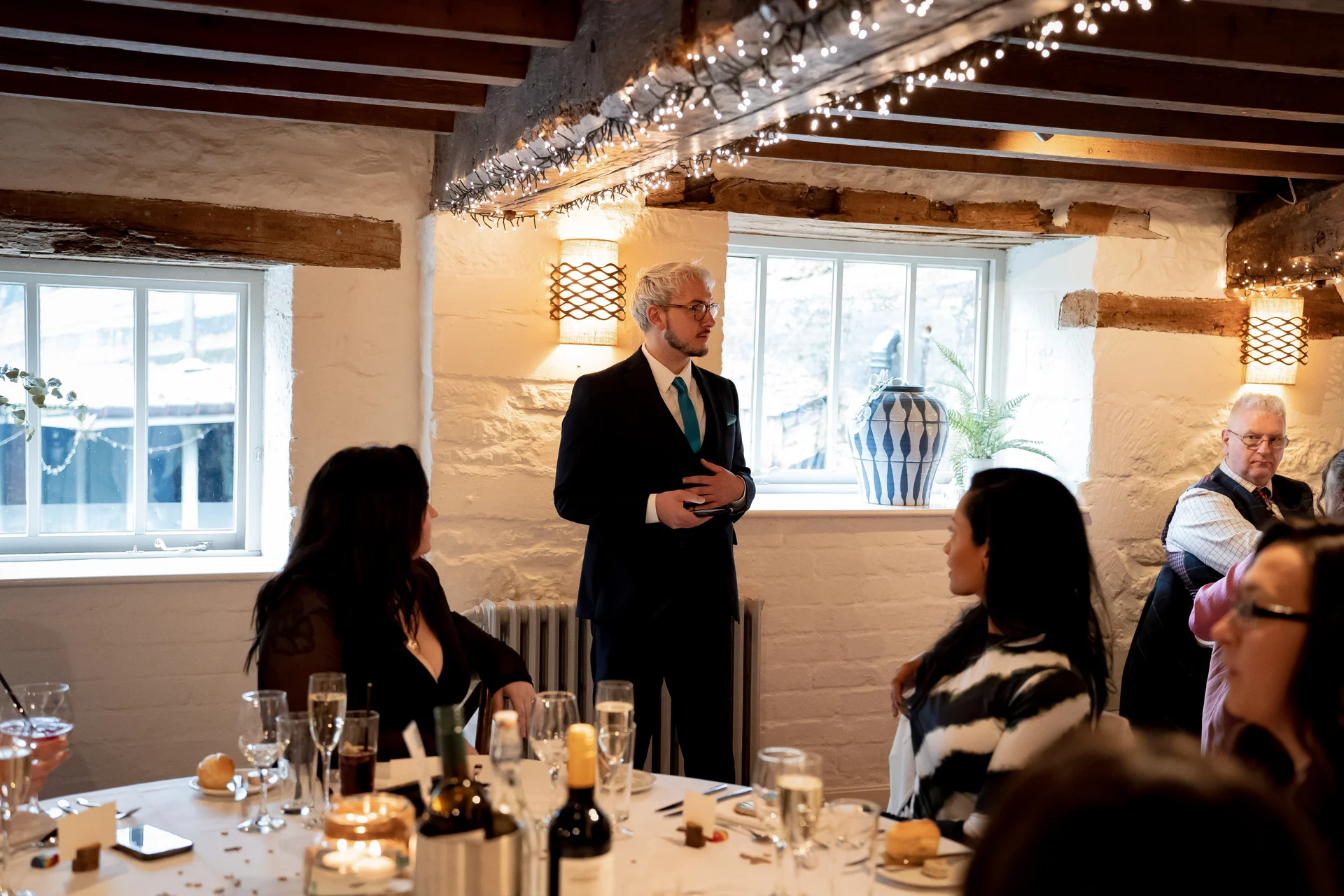 A groom delivers his wedding speech in a rustic barn venue featuring exposed wooden beams and white stone walls, as seated guests listen attentively during the wedding breakfast.