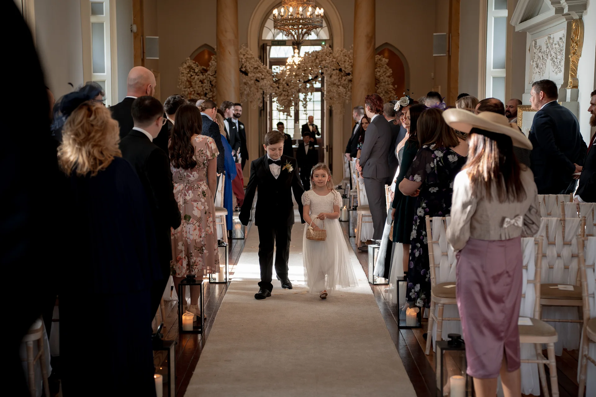 A young page boy and flower girl walk down the aisle together during a wedding ceremony inside a grand light-filled venue.