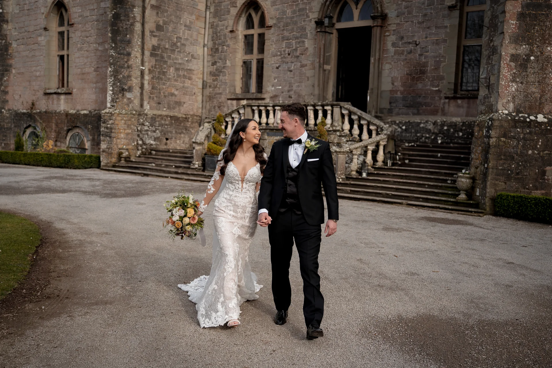 A newlywed couple happily walks hand-in-hand across a paved courtyard in front of a majestic historic stone castle, sharing a loving glance.